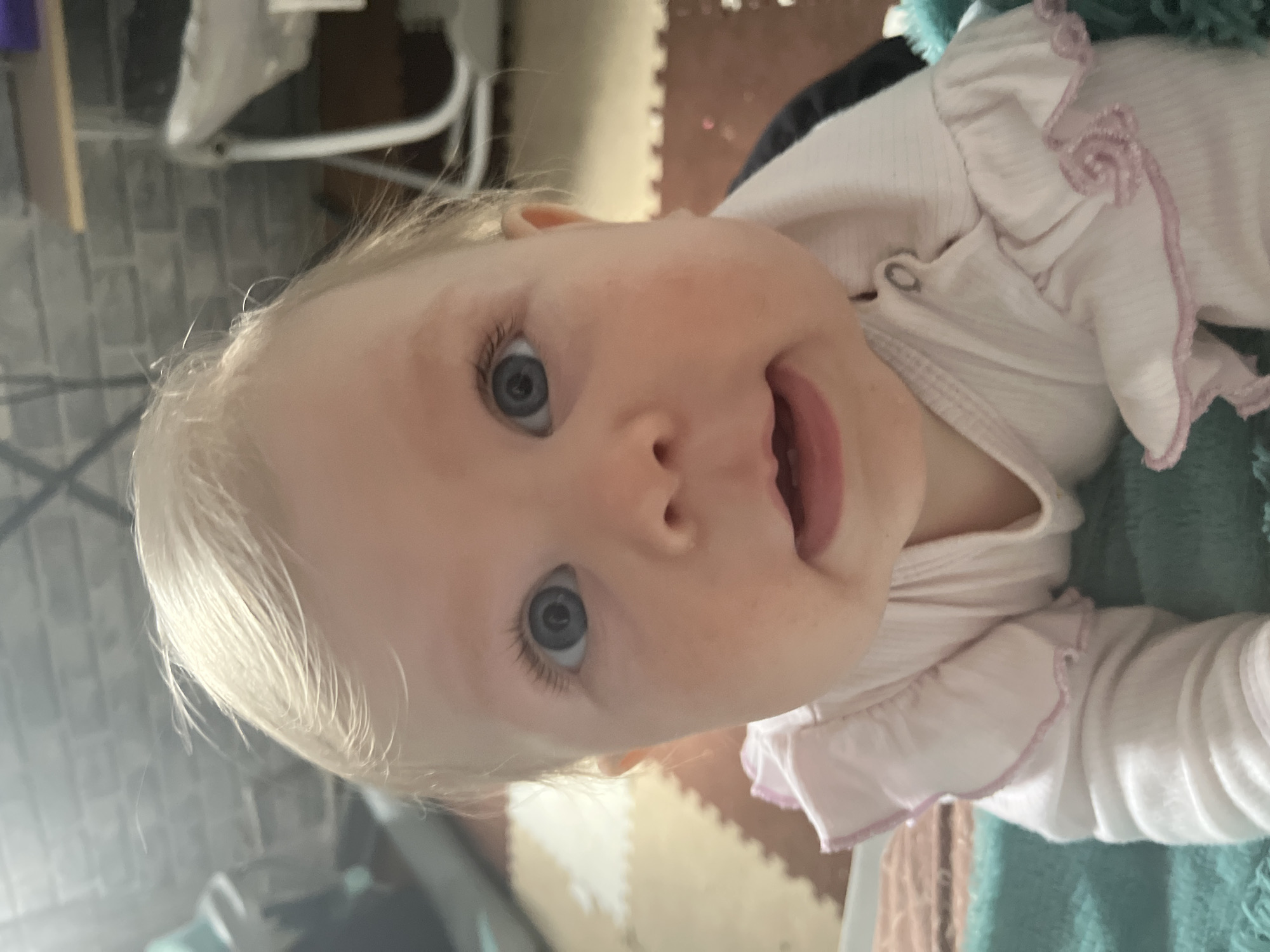 A close-up, slightly off-center portrait of a baby with fair skin, blonde hair, and blue eyes is featured in this image. The baby is wearing a light pink garment with ruffled edges. Part of the baby's mouth is open, revealing the gums and possibly the tip of the tongue. The background includes a variety of objects, including what appears to be a white metal frame, a patterned floor mat, and soft, green, textured material. No definitive elements indicate a location in Vesyoloye, Russia.