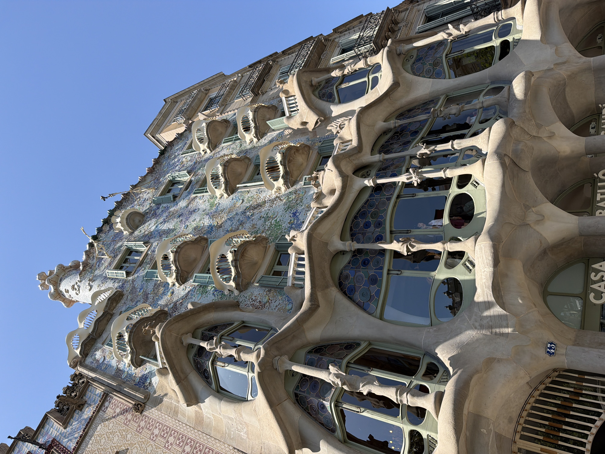 This is a low-angle, close-up shot of the Casa Batlló in Barcelona, Spain, a building designed by Antoni Gaudí. The facade features organic, flowing lines and is adorned with colorful mosaics and sculpted elements. Several balconies with bone-like shapes protrude from the building, and large, irregularly shaped windows with stained glass details are visible. The sky is clear blue and the sun casts shadows on the building. The building's unique architectural style is characteristic of Catalan Modernism.