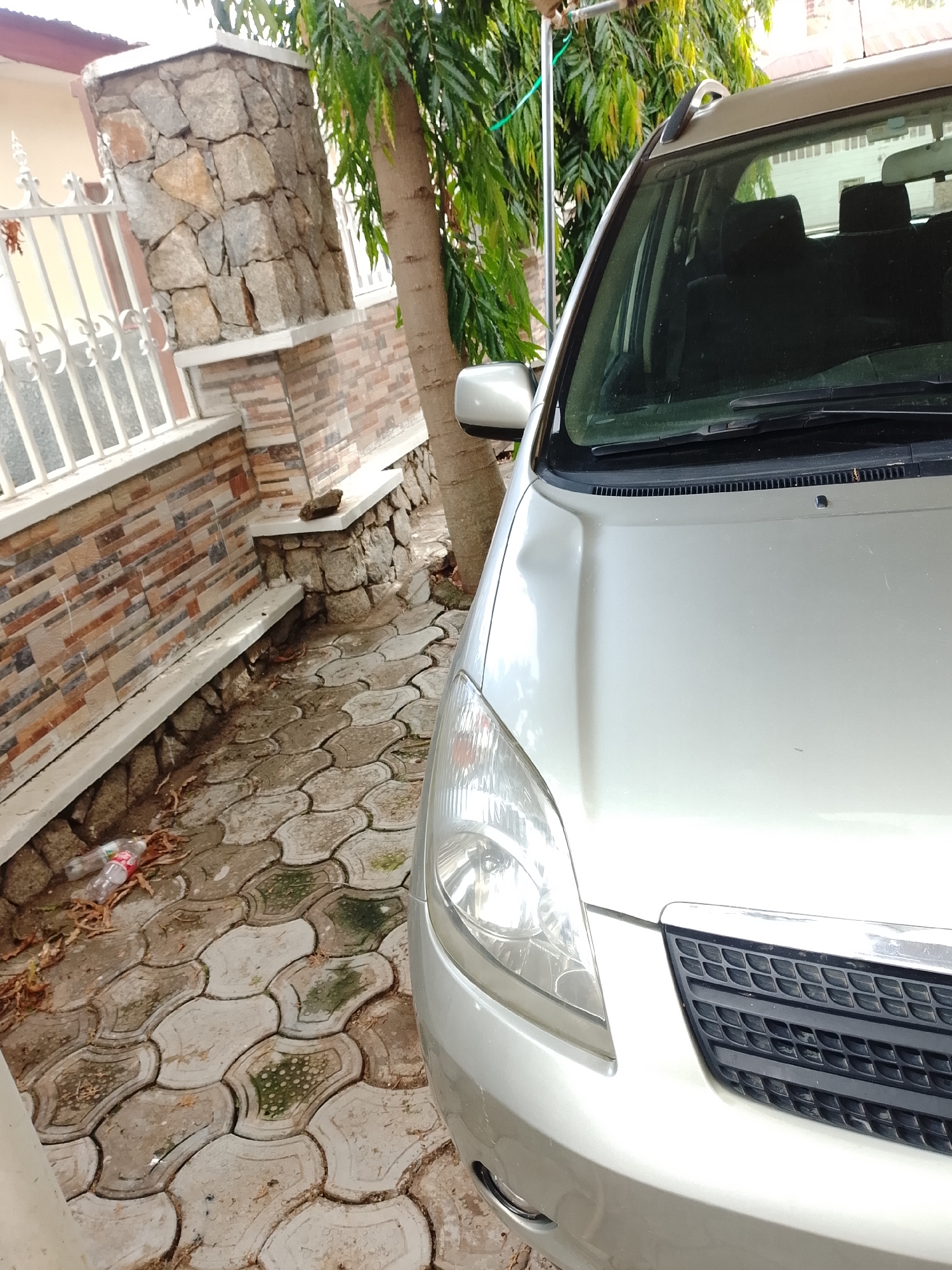 The image captures a light-colored car parked outdoors next to a stone and brick wall. The primary subject is the front right portion of a silver or champagne-colored car, likely a compact MPV or wagon, showing its headlight, hood, windshield, roof rail, and side mirror. No people are visible in the frame.
The car is stationary, with no apparent activity taking place. It is positioned on interlocking concrete pavers, which appear wet in sections and show patches of green moss or algae growth, suggesting damp conditions or recent rain. Scattered dried leaves are also on the ground.
Notable details include a sturdy wall and fence structure to the left of the car. This structure features a pillar built from irregularly shaped natural stones, with a capping stone at the top. The wall extending from it is made of rectangular, multi-toned bricks in shades of red, brown, and tan. A white, ornate metal fence runs along the top of this wall. A large tree with abundant green, drooping leaves stands behind the car and the wall, its trunk visible. The lighting suggests it is daytime, with bright but diffused light, indicating either an overcast sky or indirect sunlight. A red and white plastic bottle, too blurry to read any text, is seen on the ground near the base of the wall. The car's hood has a minor indentation or reflection near the windshield.