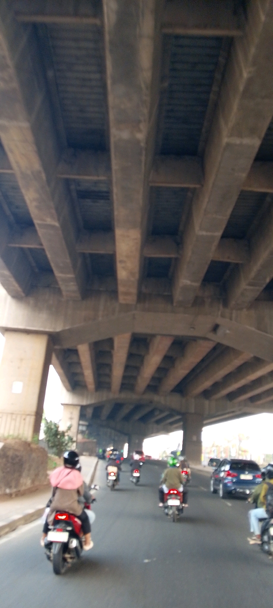 The image depicts a street scene in Bekasi, Indonesia, under an elevated highway structure. Several motorcycles are visible on the road, with riders wearing helmets and some carrying passengers. One motorcycle in the foreground has a rider in a pink headscarf and a tan jacket, and another passenger behind them. Another motorcycle further ahead has a rider in a green jacket and helmet. A blue car is also present on the road. The underside of the highway is made of concrete beams and supports, creating a covered passage for the traffic below. The lighting suggests it is daytime.