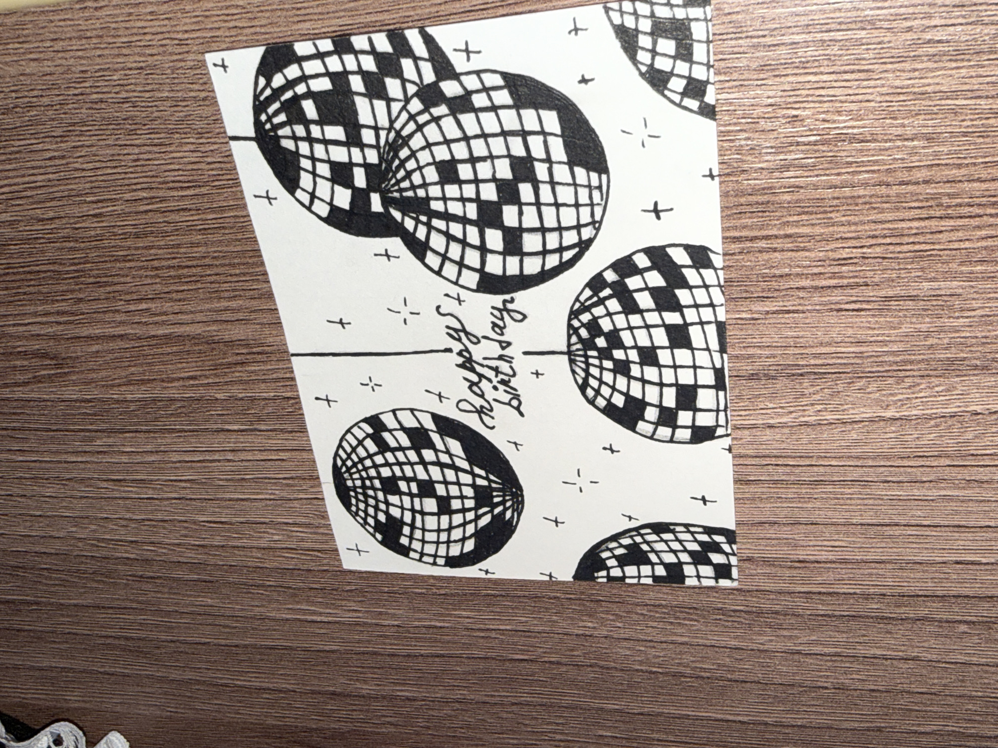 The image shows a handmade birthday card with a drawing of disco balls and the words "Happy Birthday" written in cursive. The card is placed on a wooden surface. There is no information about the location in Rayevskiy, Russia. The drawing style is simple, with black ink on a white background. The disco balls are depicted with a grid pattern, representing their reflective surfaces. Some small crosses and asterisk-like shapes are scattered around the disco balls, adding to the festive theme. The wooden surface appears to have a natural grain pattern and a warm brown color. The lighting in the image suggests an indoor setting.