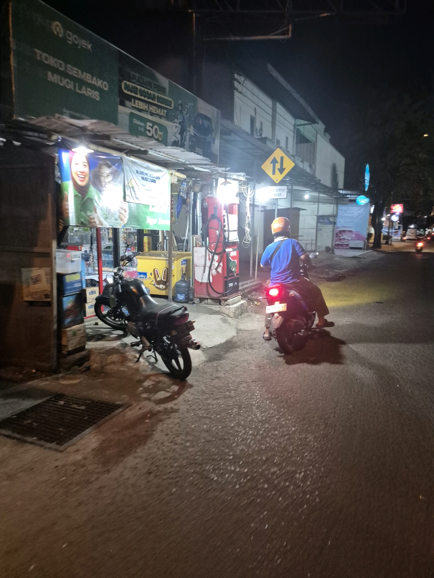 The image depicts a night scene on a paved street in Surakarta, Indonesia. In the foreground, a black motorcycle is parked on the left, with a partial view of a gas pump to its right. A person wearing a blue shirt and an orange helmet is riding a motorcycle away from the viewer on the right side of the street. The rider's motorcycle's taillight is illuminated. In the background, a business with green signage and advertisements can be seen. The street is illuminated by artificial lights, casting shadows on the ground. There are other businesses and traffic lights visible further down the street.