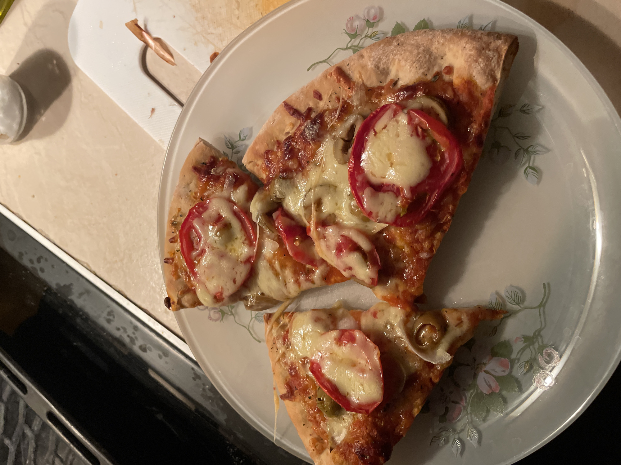 Three slices of pizza are arranged on a light-colored, floral-patterned plate. The pizza slices appear to have a tomato sauce base, melted cheese, sliced tomatoes, and possibly some green peppers and olives. The crust is golden brown and appears to be slightly charred in places. The plate is situated on a countertop that is a light beige or cream color with a subtle pattern. To the left of the plate is the edge of a dark appliance, possibly a stove or oven, with a textured surface. In the upper right corner of the image, a white cutting board with a few small pieces of food and a small piece of what looks like a carrot are visible. A small glass jar containing white salt is also partially visible in the background. The lighting suggests an indoor setting, possibly a kitchen. The scene is devoid of any people or clear indicators of the specific location within Zlatograd, Bulgaria, beyond it being an indoor domestic setting.