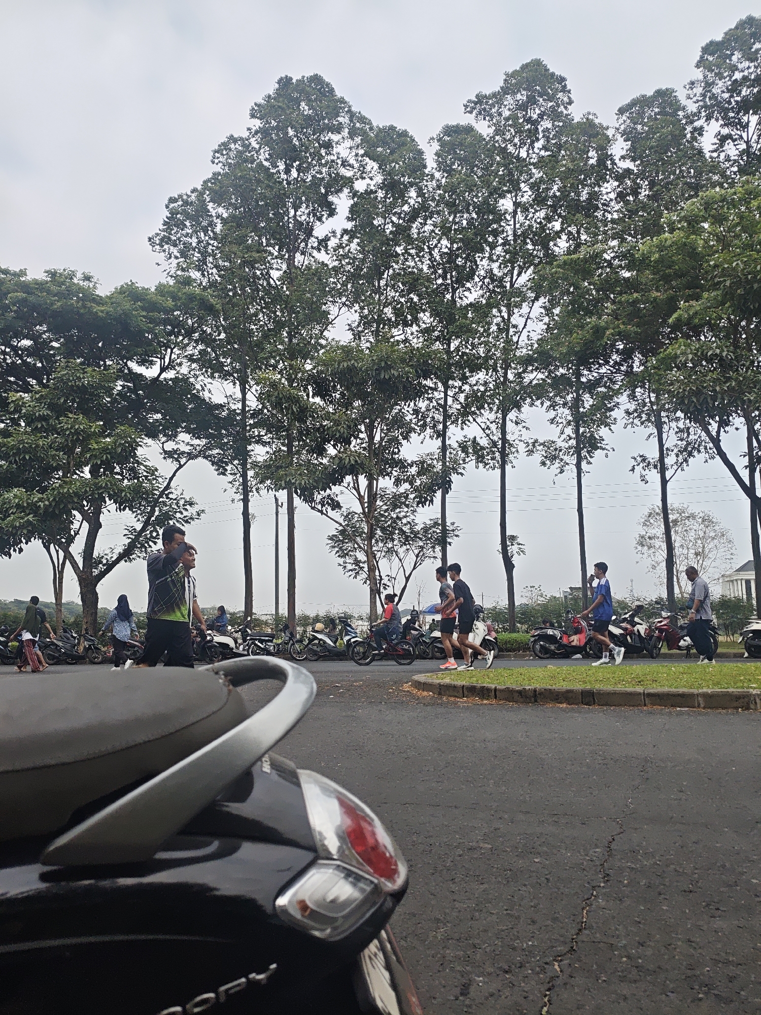The image depicts an outdoor scene in the city of Bacin, Indonesia, with a focus on a paved road bordered by a grassy median and tall trees. In the foreground, a portion of a black scooter is visible. The midground features a paved area with numerous parked motorcycles, and several individuals are walking and jogging. A group of three young men are jogging together, two in athletic attire and one in a blue shirt and white shorts. Another man, wearing a gray shirt and black pants, is seen walking. To the left, two people are walking towards the parked motorcycles, one wearing a green shirt and the other a blue and white patterned shirt. Further in the background, more parked motorcycles and a few individuals are present, along with buildings and more trees. The sky is overcast.