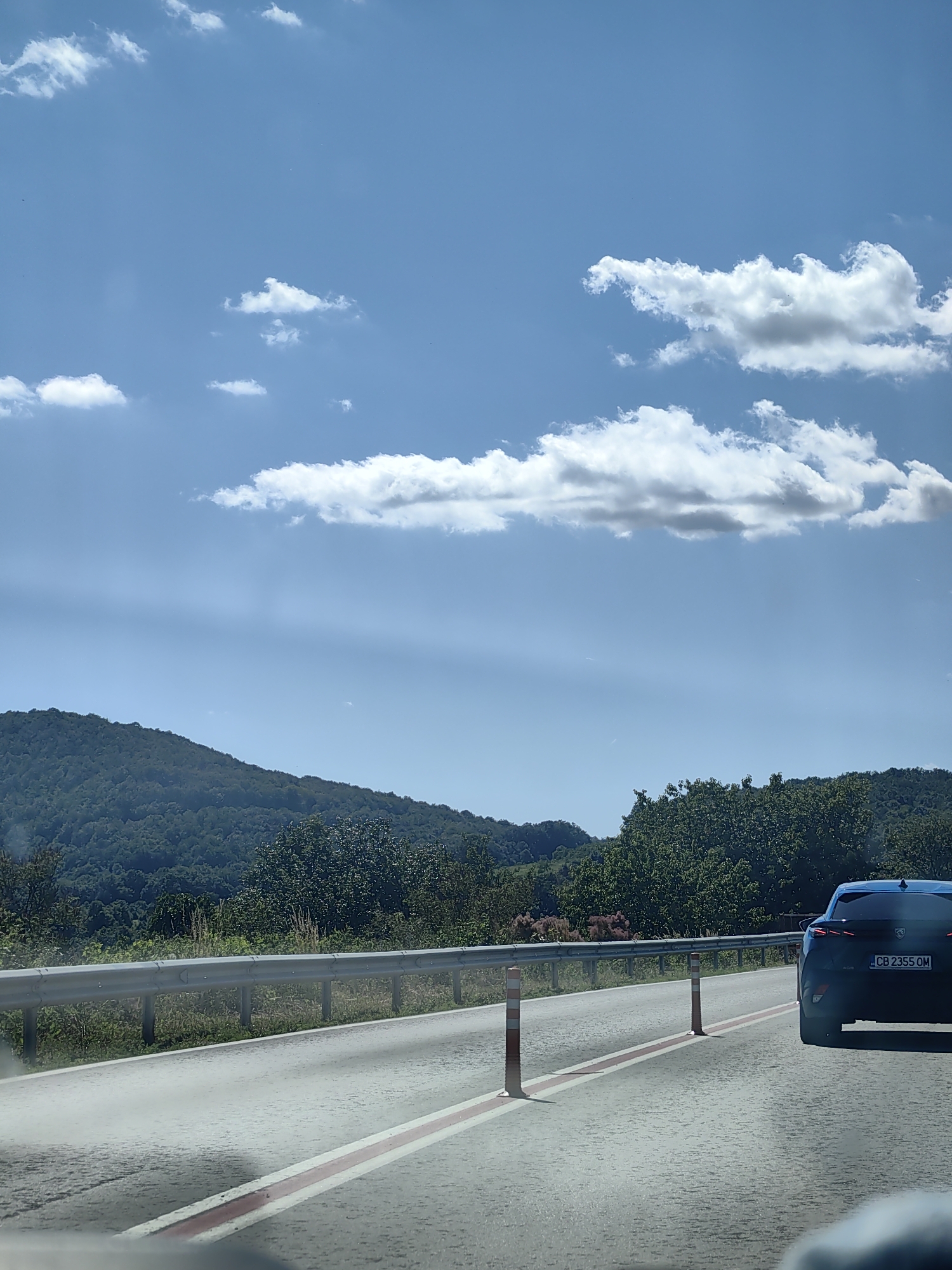 The image depicts a scenic view from a vehicle traveling on a road near Stefanovo, Bulgaria. The road is paved with visible red and white lane markings. A silver guardrail runs alongside the road, separating it from a grassy verge. Beyond the verge, a densely forested hillside rises, covered in lush green trees. In the distance, rolling hills are visible under a bright blue sky dotted with fluffy white clouds. A dark blue car with license plate "CB 2355 OM" is visible on the right side of the road, traveling in the same direction as the viewer. Orange and white traffic cones are placed at intervals on the side of the road. The overall impression is one of a clear, sunny day in a rural, mountainous region.