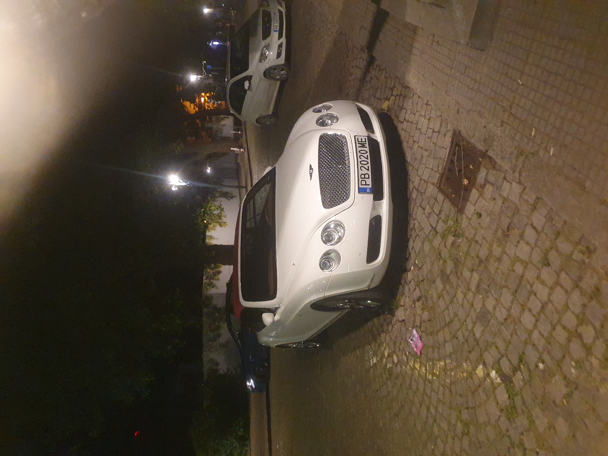 The image shows a white Bentley Continental with a Bulgarian license plate (PB 2020 ME) parked on a cobblestone street at night in Plovdiv, Bulgaria. A second white car is visible behind the Bentley. Street lights illuminate a building in the distance. The ground shows a patch of light-colored cobblestone with some litter. The background has trees and what appears to be a tall wall.