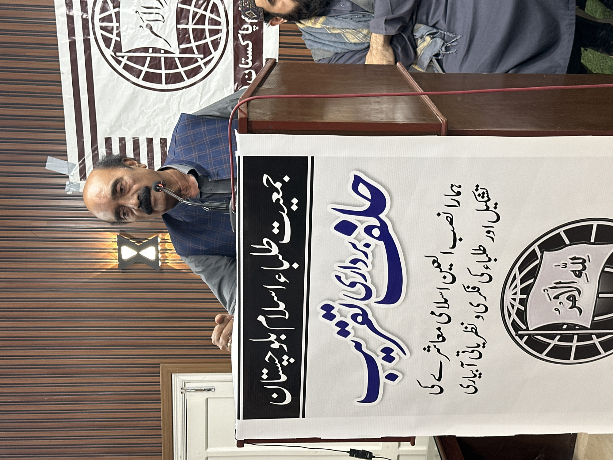 A man is speaking at a podium in Quetta City Tehsil, Pakistan. The podium has a sign that says "Jamiat Tulaba Islam Balochistan" and "Oath-taking ceremony." There are two other people in the background. The wall behind the man is striped.