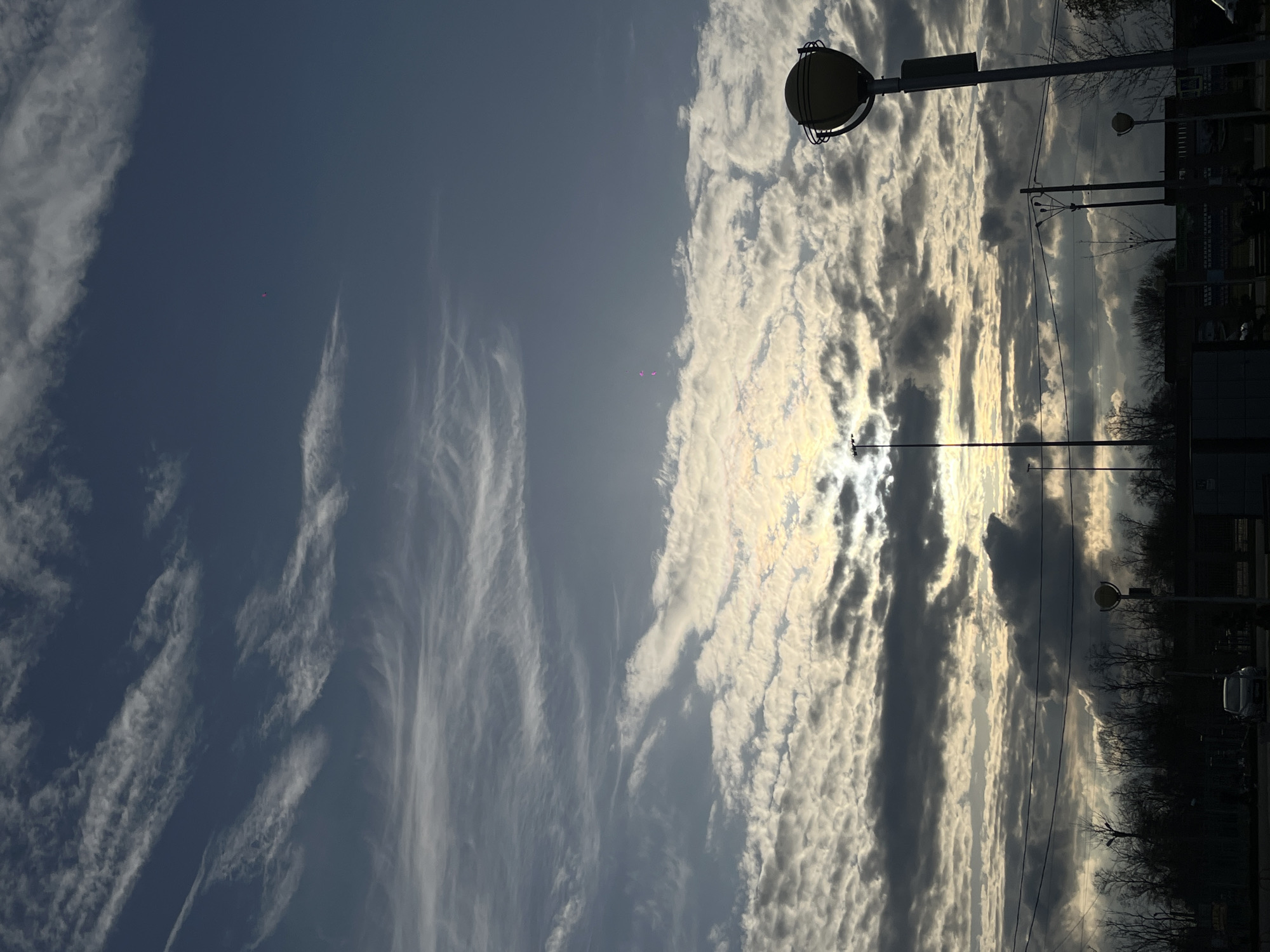 The image shows a sky with a mix of cirrus clouds on the left side and cumulus clouds on the right, with the sun shining through. On the right side of the image, there are silhouettes of trees, buildings, and streetlights. There is no information to confirm the location as Novopokrovskaya, Russia. There are no people present in the image.