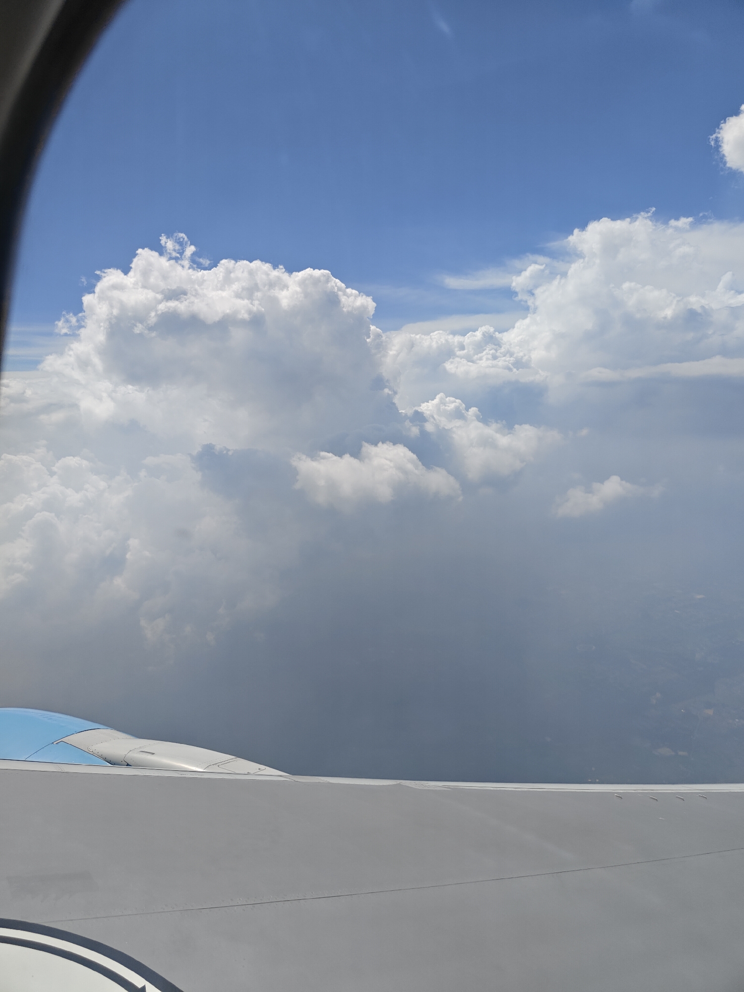 This image, captured from an airplane window, depicts a vast expanse of the sky filled with dramatic cumulus clouds. The clouds are predominantly white, with some areas showing darker, gray undertones, suggesting varying densities and perhaps approaching weather. The sky above is a clear, bright blue, indicating a sunny day. In the foreground, a portion of the airplane wing is visible, its surface a smooth, light gray. A blue and white section of the aircraft's engine or fuselage protrudes slightly above the wing. Far below, through a haze created by the distance and atmospheric conditions, faint outlines of land and possibly man-made structures can be discerned. The image does not contain any people or any specific identifiable landmarks within Kahaksona, India, or any other location.