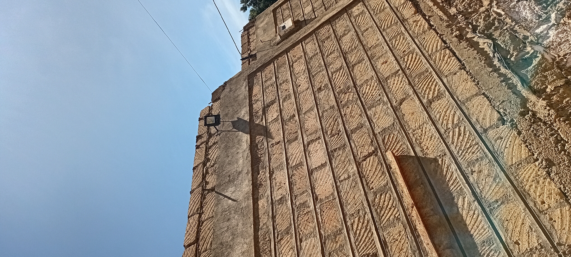 The image shows a close-up view of a tiled roof of a building under construction or repair, set against a clear blue sky. The tiles are reddish-brown and have a textured, possibly handmade, appearance. They are laid in horizontal rows, with a distinct pattern of grooves or indentations on each tile, creating a visual texture. Thin, dark lines, possibly grout or embedded reinforcement, run horizontally between some of the rows of tiles.
The roof appears to be sloped, with a ridge line visible at the top of the frame. A black, rectangular floodlight is mounted on a bracket on the roof near the ridge, casting a long shadow. Further along the ridge, a small white electrical box or meter is visible. There are power lines visible against the sky. The edges of the roof show rough concrete work and what appears to be construction debris.
The setting is outdoors during the day, indicated by the bright sunlight and the clear sky. The overall impression is of a building undergoing work, possibly a residential or community structure in a semi-urban or rural area. The location is specified as Kijabe ward, Kenya.