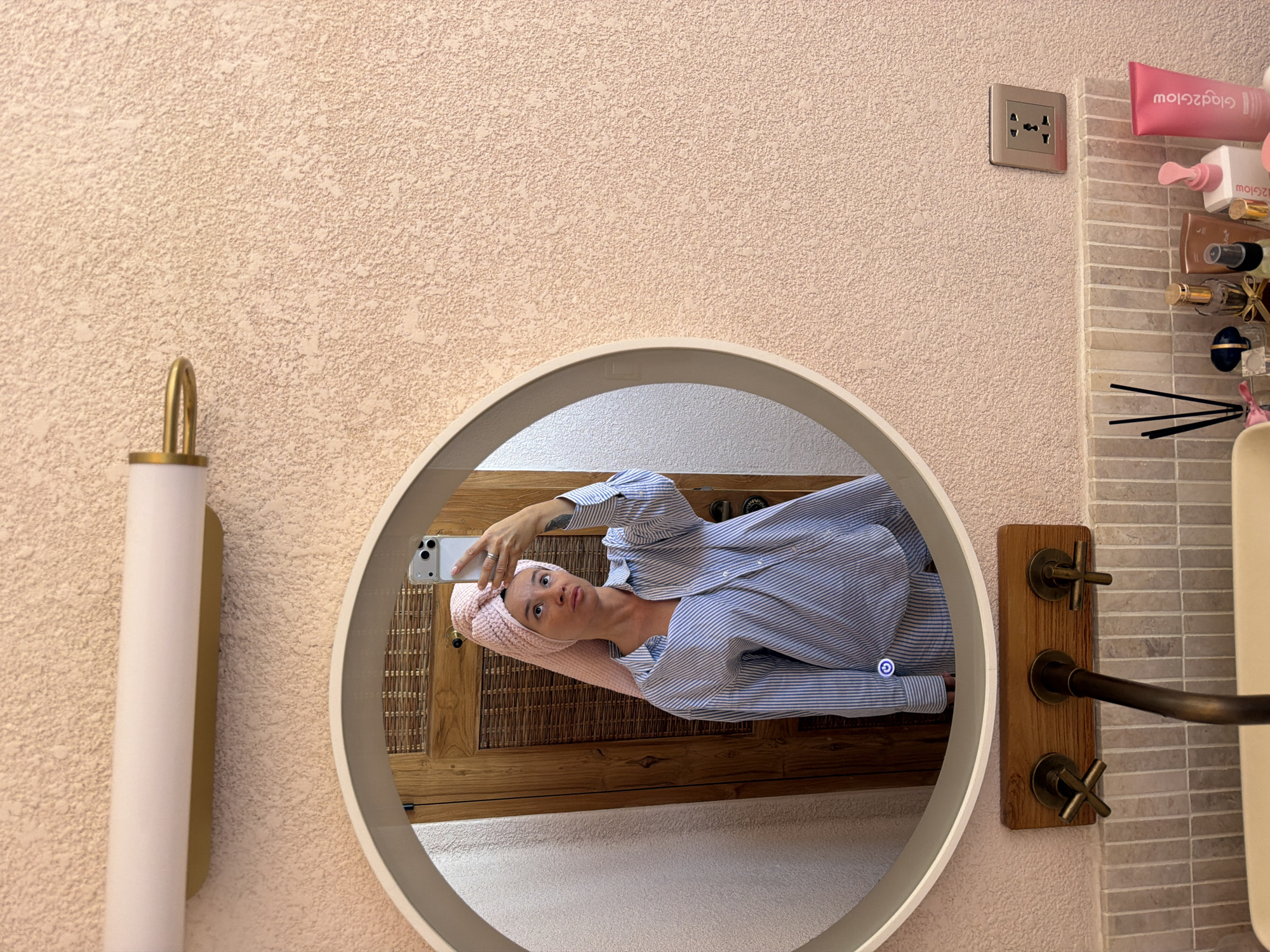 A woman with a pink towel wrapped around her head takes a selfie in a bathroom mirror. She is wearing a blue and white striped oversized shirt. The bathroom has a textured pink wall and a round mirror with a built-in light. Below the mirror are a wooden panel with three faucets and a tiled backsplash. On the counter, several beauty products and diffusers are visible, including a pink tube labeled "Glad2Glow". An electrical outlet is mounted on the wall to the right. The overall scene suggests a domestic setting, likely a personal bathroom. The specific location of Mengwi, Indonesia, is not depicted visually.