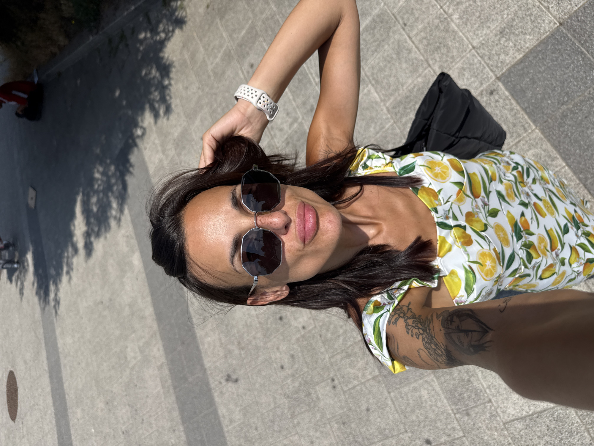 A woman is taking a selfie outdoors on a sunny day. She has dark hair, is wearing sunglasses, and a white dress with a lemon print. She is also wearing a white fitness tracker on her left wrist. A black bag is slung over her shoulder. The background is a paved area with some shadows from trees. In the distance, a person in a red shirt is sitting on a curb. The scene appears to be in a public outdoor space, possibly a park or promenade. The city of Pleven, Bulgaria, is not identifiable from the visual information provided in the image.