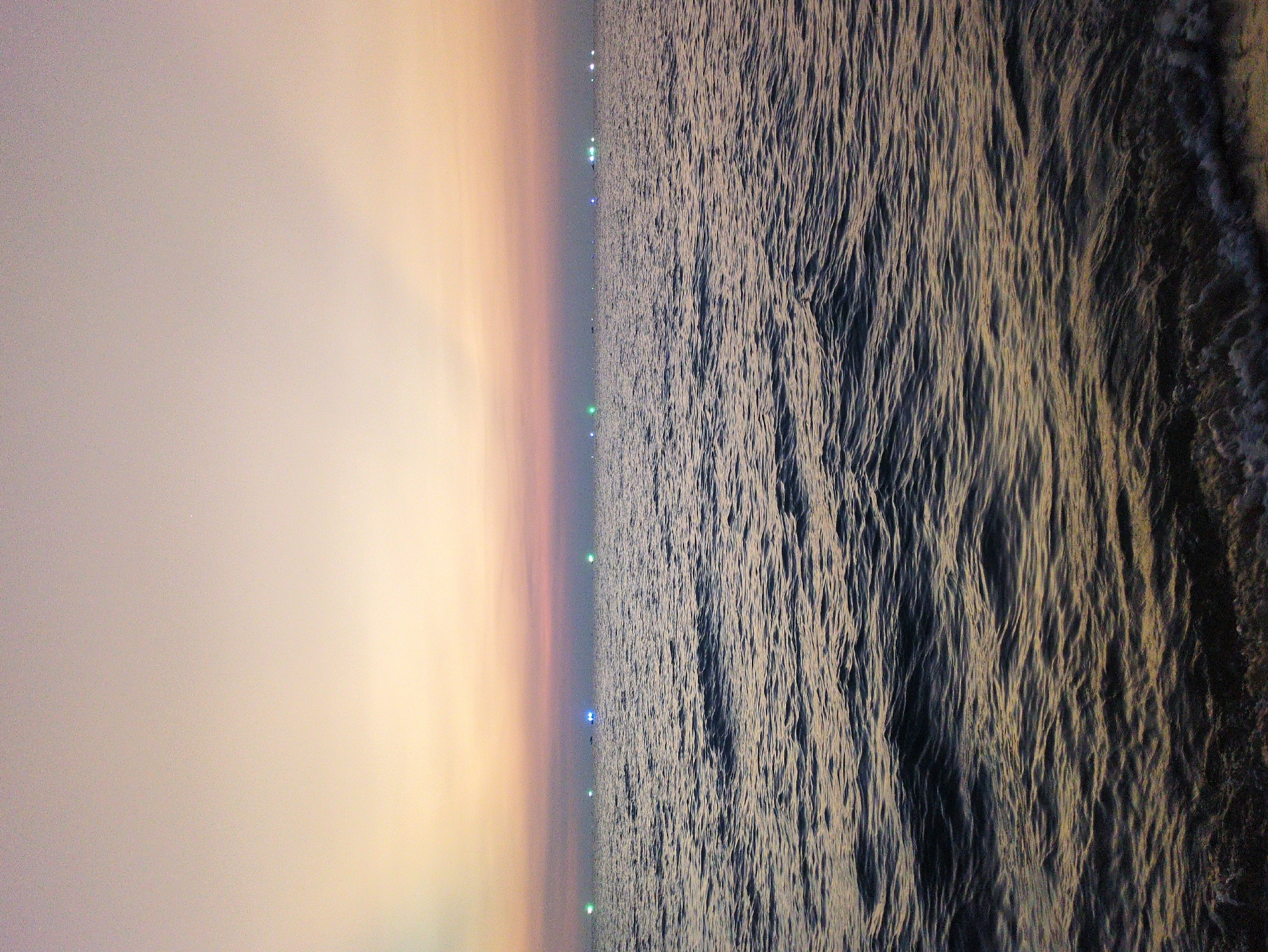 This image shows a nighttime ocean scene. The sky on the left side of the image is a blend of light pink and orange. A line of spaced, small lights runs horizontally across the middle of the image. The right side of the image depicts the ocean, its surface reflecting the light and appearing textured with waves. This is the sea in the city of Phú Quốc, Vietnam.