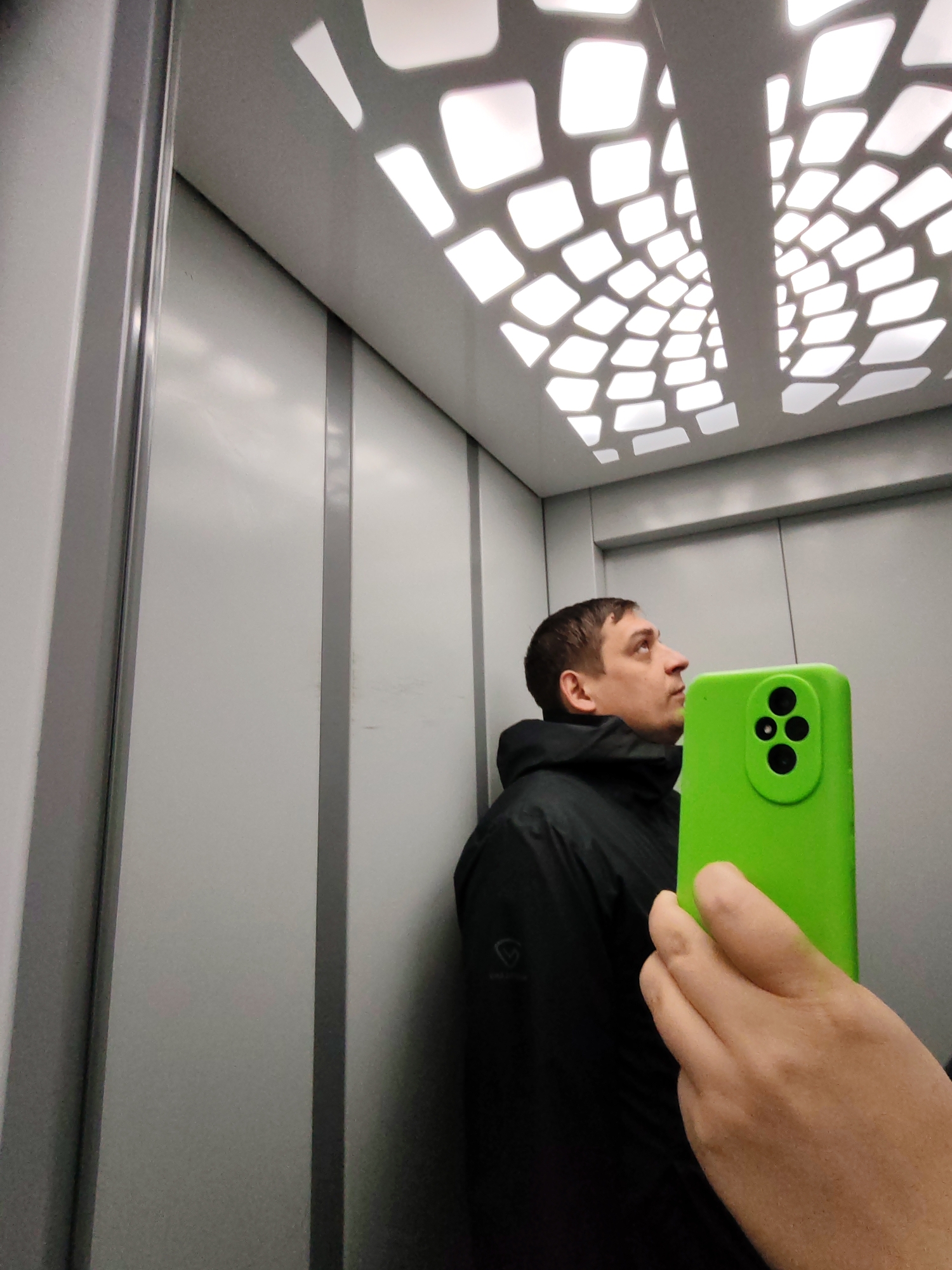 The image captures a man taking a selfie within what appears to be a modern elevator, captured via a reflection. The man, seen from the chest up and in profile, is looking upwards and slightly to the right. He has short, dark brown hair and a neutral expression. He is wearing a dark, possibly black, hooded jacket that features a faint, partially obscured logo on the left chest area, which includes letters such as "PRO".
In the foreground, a hand holds a bright neon green smartphone, positioned to partially obscure the man's face. The rear of the phone is visible, showcasing its black camera lenses (three prominent ones and possibly a smaller fourth or a flash).
The setting is an elevator interior, identified as being in Podolsk, Russia, per the provided context. The walls are composed of light gray or off-white panels segmented by vertical metallic or dark gray strips. The most distinctive feature of the elevator is its ceiling, which boasts a stylized lighting design. It comprises numerous illuminated, geometric quadrilateral shapes (squares and rectangles with rounded corners) arranged in a grid-like pattern that appears to recede towards the center, creating a sense of depth and contemporary aesthetics.
The man's action of looking upwards suggests he might be admiring the unique ceiling design or simply posing for the reflected selfie. The lighting within the elevator is bright, originating from the ceiling fixture. No specific time of day or external weather conditions are discernible.