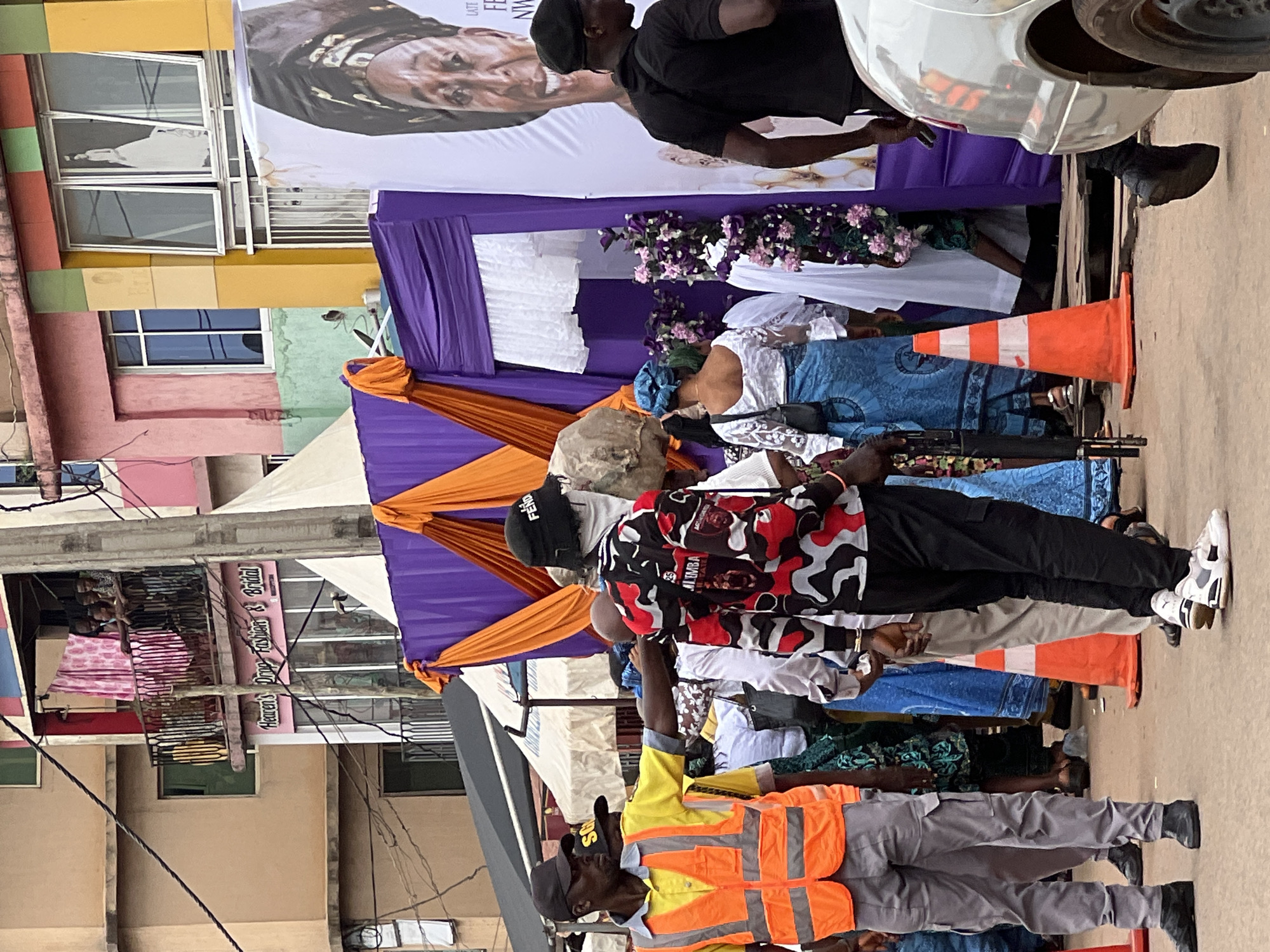 The image depicts a street scene in Nkpor, Nigeria, likely during a public event or gathering. In the foreground, a security guard in a high-visibility orange vest and grey pants is directing people. To his right, a person in a camouflage-patterned jacket is standing, facing away from the camera. Behind them, a large banner features a portrait of an older woman. The background shows buildings with colorful facades and balconies, with some people observing from above. Several orange traffic cones are strategically placed, suggesting crowd control or the delineation of an area. Purple and orange drapes are set up, possibly as decoration for an event. The ground appears to be a paved street, and a silver car is visible on the right side of the frame. The overall atmosphere suggests a busy, community-oriented event.