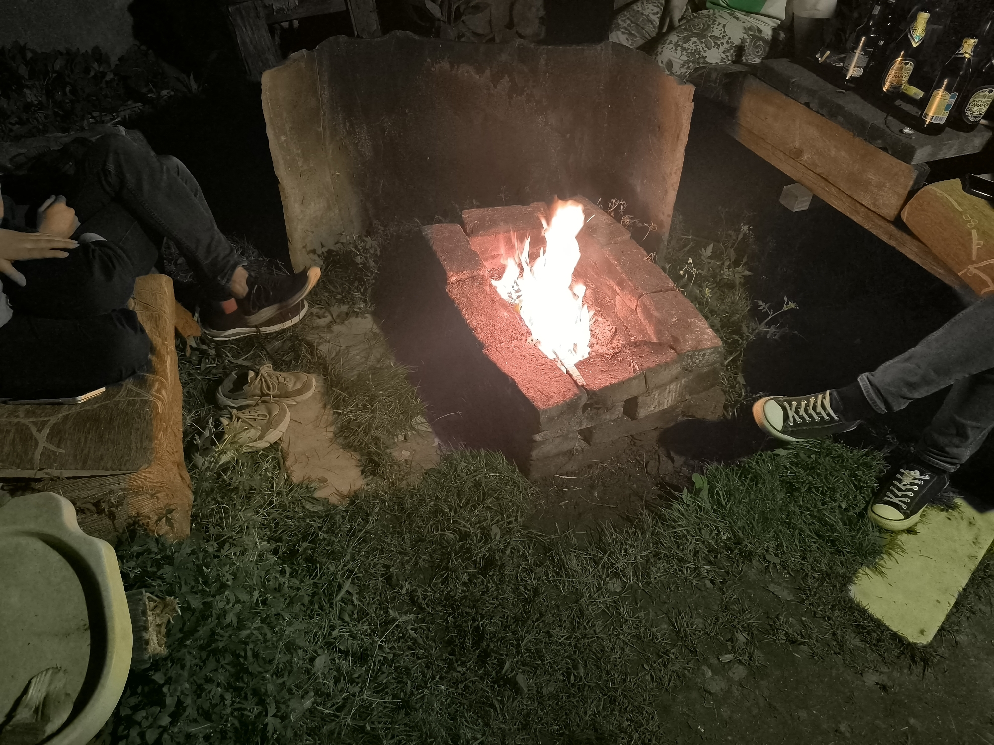 The image depicts an outdoor scene at night, likely a backyard or recreational area, with a small bonfire in a brick fire pit as the central focus. Several individuals are present, with their legs and feet visible, suggesting they are sitting or relaxing around the fire. One person on the left is wearing dark jeans and sneakers, with a hand resting on their lap. Another person on the right is wearing dark pants and black sneakers with white laces and yellow soles. There are also shoes scattered on the ground near the fire pit. The environment includes grass, dirt, and a wooden bench. In the background, there are some bottles on a wooden surface and a rough-hewn stone structure behind the fire pit. The lighting is dim, with the fire providing the primary illumination. The location is identified as the city of Кашино, Russia.
