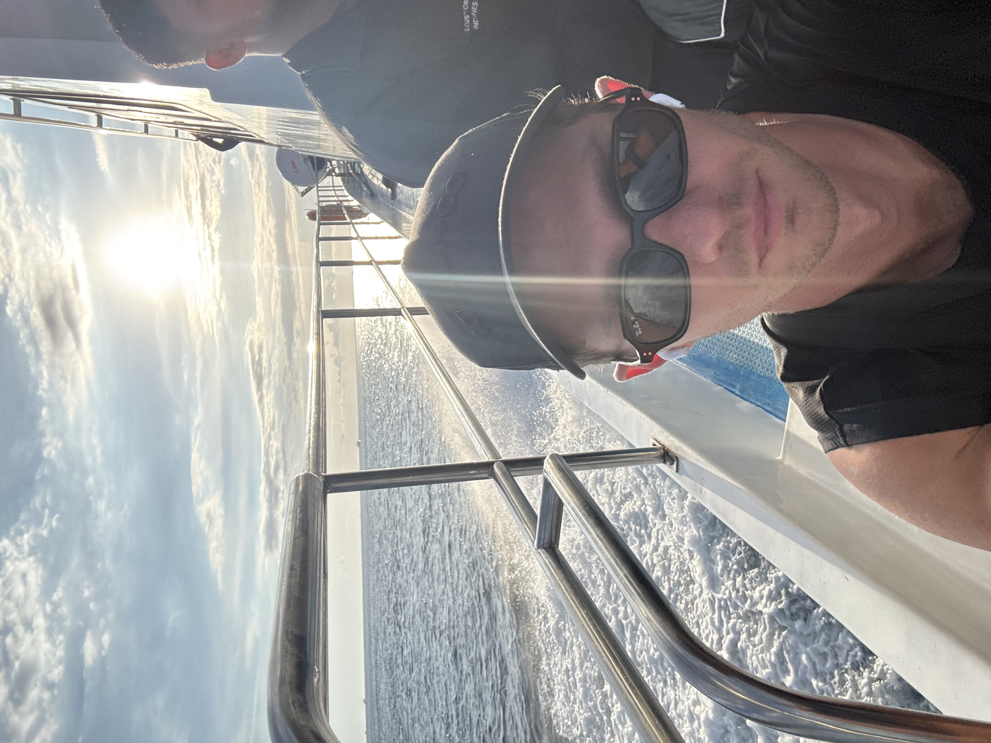 A portrait-oriented photo captures a man taking a selfie on a moving boat under a bright, low-lying sun. The man, in the foreground, wears a dark baseball cap with a faint logo (possibly "GQ") and dark Ray-Ban sunglasses, and a dark t-shirt. He has a trimmed beard and mustache and a relaxed, slight smile. The sun, prominent in the upper left, creates a strong glare across the sky and the water. Behind him, another person is partially visible, wearing a dark shirt with text that appears to read "LOUIS VUITTON" and "INDONESIA" in separate lines.
The boat's chrome railings are visible in the foreground and midground. On the right side of the frame, white foam and water spray are clearly visible, indicating the boat is moving at speed through the water. In the midground, a white satellite dish, possibly marked "CSM," is mounted on the vessel. The sky is bright with scattered white clouds. The overall scene depicts a sunny day, likely during morning or late afternoon, with a relaxed atmosphere on open water.