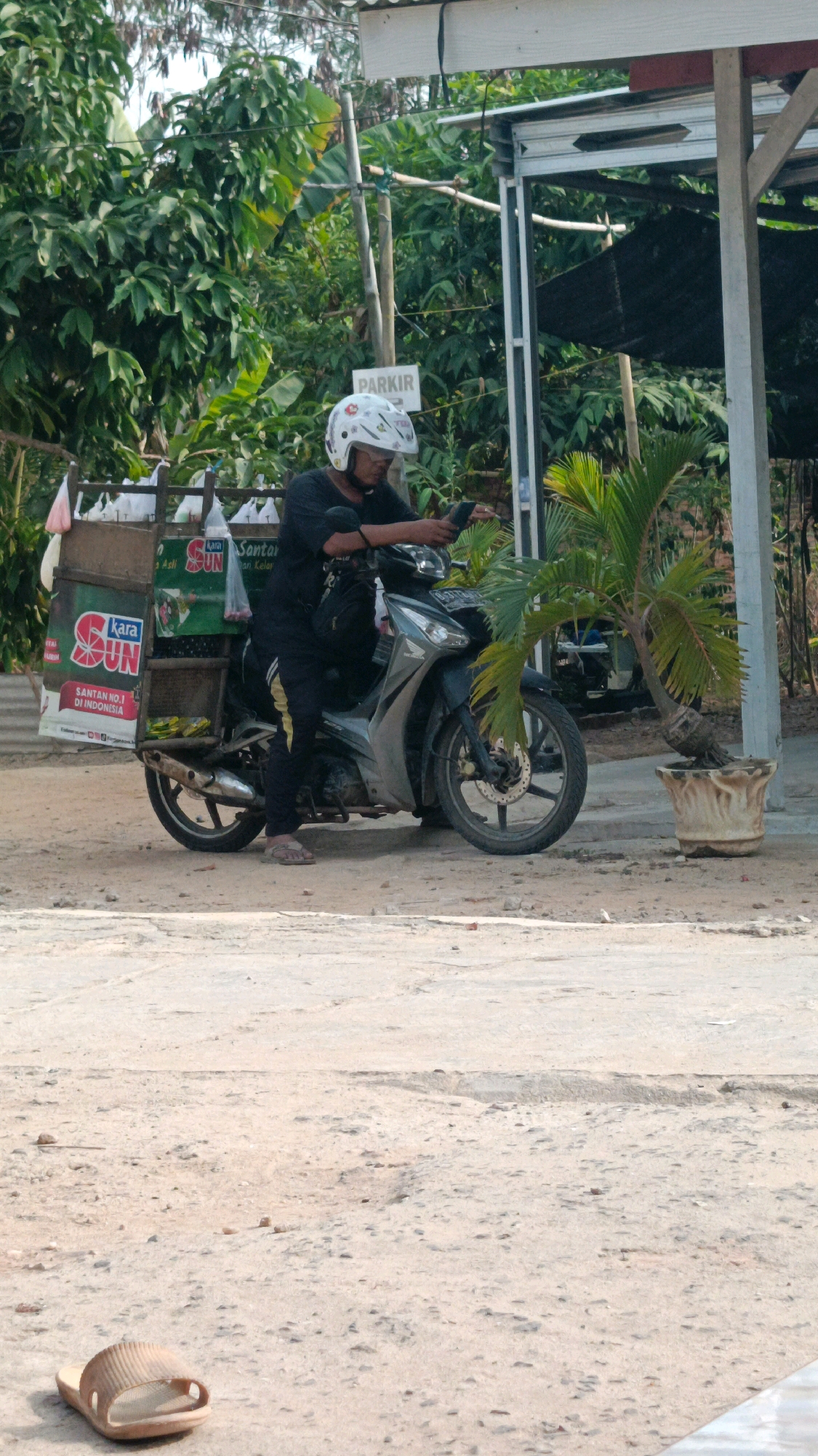 A man wearing a white helmet and black clothing is sitting on a gray motorcycle with a delivery cart attached to the back. He is looking at his phone. The motorcycle is parked on a dirt and concrete area. In the background, there are trees and a structure with a "PARKIR" (parking) sign. A potted palm tree is to the right of the motorcycle. The scene appears to be in Metro, Indonesia, based on the visible product branding, which includes "KARA SUN".