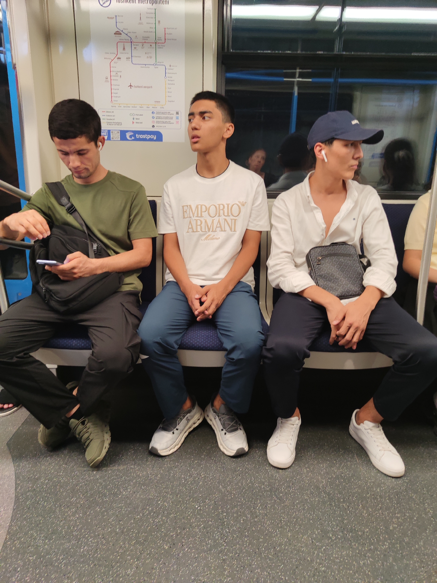 Three young men are seated next to each other on a subway train. The man on the left is wearing a dark green t-shirt and black pants, and is looking down at his phone. He has a black crossbody bag over his shoulder. The man in the middle is wearing a white t-shirt with a graphic print and blue pants, and is looking straight ahead with his hands clasped. The man on the right is wearing a white button-down shirt, dark blue pants, and a navy blue baseball cap. He is looking to his right and has a small patterned crossbody bag. All three men are wearing sneakers. Behind them, a subway map is visible on the wall. The seats are blue and the floor is a speckled gray. Reflections of other passengers are visible in the window.