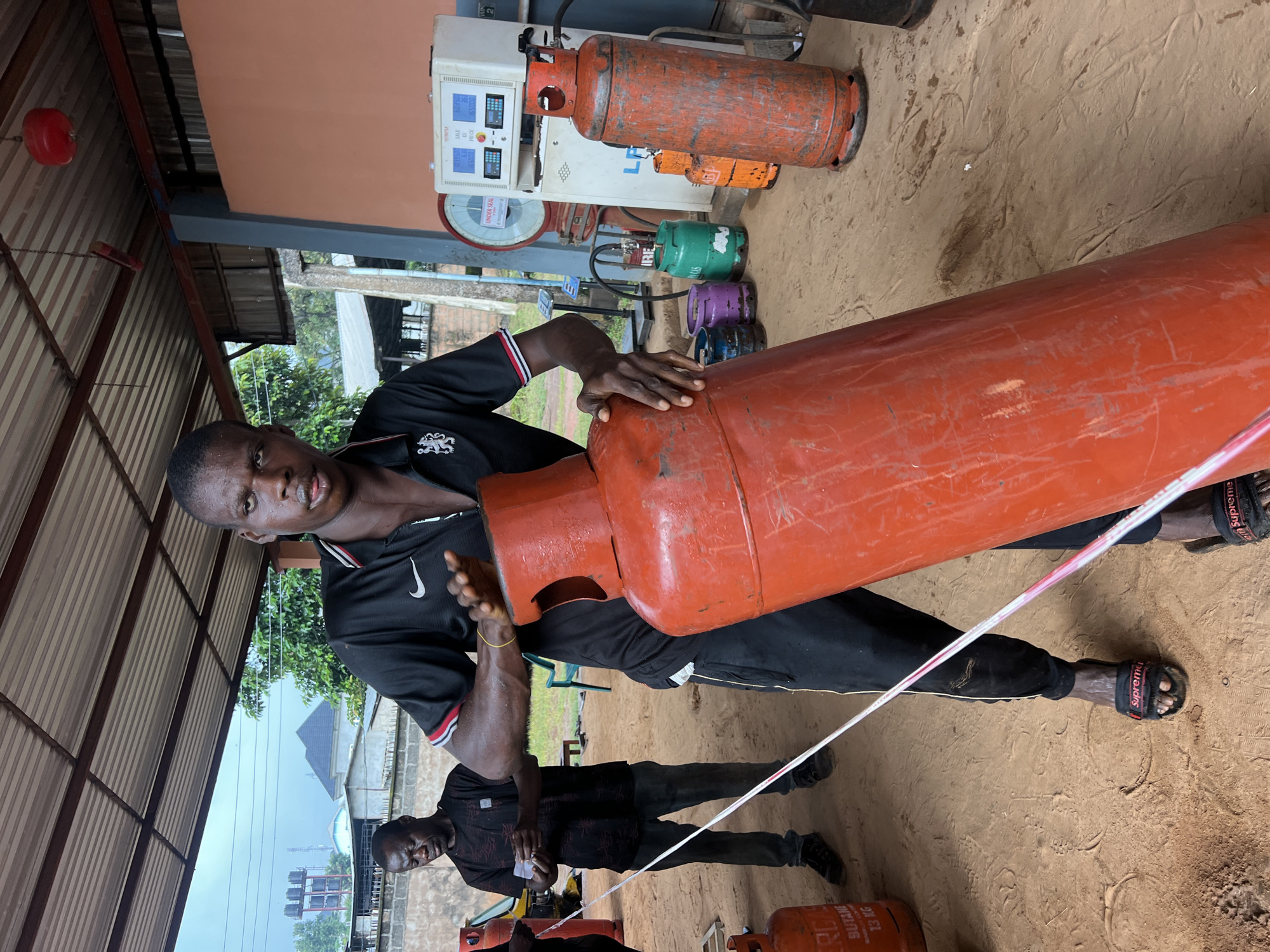 In Nkpor, Nigeria, a man is shown holding a large orange gas cylinder. He is wearing a black t-shirt with a white logo and black pants. Another man is standing behind him, also in dark clothing. The scene appears to be an outdoor gas filling station, with a gas pump and several other smaller gas cylinders in the background, some colored green, purple, and orange. The ground is sandy and appears to be a dirt lot. The overall impression is one of a busy day at a local business, with the focus on the cylinder being handled. The man in the foreground has a determined expression, suggesting he is engaged in a task that requires strength and concentration. The presence of the gas pump and various cylinders indicates a commercial setting where fuel is dispensed. The time of day appears to be daytime, likely afternoon given the lighting. The weather seems to be overcast or hazy, as the sky is bright but not intensely sunny. There is text visible on some of the gas cylinders, including "BUTANE 13 KG". The scene conveys a sense of everyday life and commerce in the Nigerian setting.