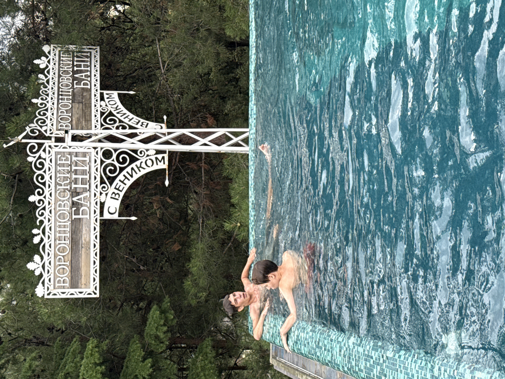 In this image, two young males are submerged in a turquoise swimming pool with a mosaic tiled edge. One individual, with light brown hair, leans on the pool's edge, his arms resting on the surface. His expression is somewhat strained or surprised. The other, with darker brown hair, is more submerged, with his back to the camera, also resting his arms on the edge. A third person is partially visible in the water. Behind them, a white metal sign with ornate scrollwork is prominently displayed. The sign features Russian text, with "ВОРОНЦОВСКИЕ БАНИ" (Vorontsovskie Baths) visible on two panels, along with "С ВЕНИКОМ" (With a broom) on a lower curved panel. The background consists of dense green pine trees. The overall impression is a recreational scene at a location identified as Vorontsovskie Baths, likely within the city of Yalta, Ukraine, given the Russian text on the sign.