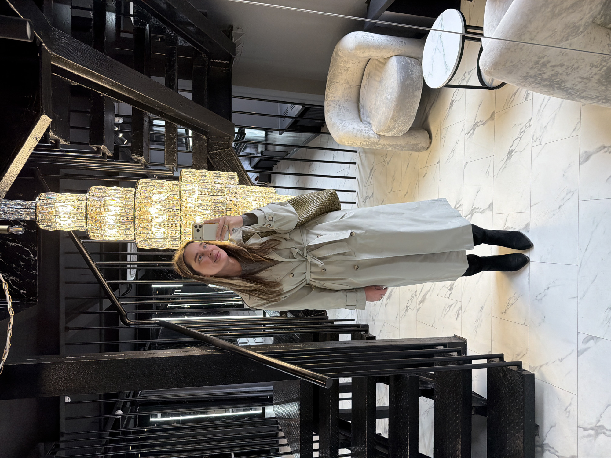 The image shows a woman taking a selfie in an interior space with modern architectural elements. She is standing on marble-tiled flooring next to a black metal staircase with open risers. The woman is wearing a light-colored trench coat and black boots, holding a phone in her left hand. Above her hangs a multi-tiered crystal chandelier. In the background, there are modern chairs and a small, round side table. The scene suggests a stylish or upscale environment, possibly a hotel or boutique. The location is the Ritz-Carlton Hotel in Moscow, Russia.