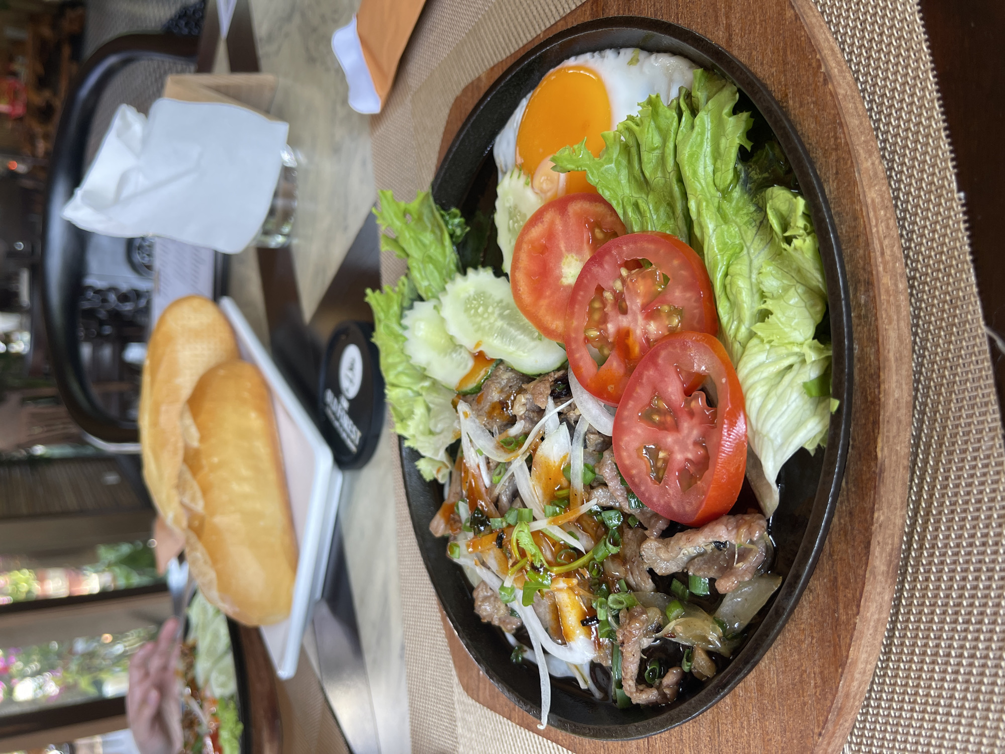 The image displays a vibrant meal served in what appears to be a restaurant or cafe setting in Phú Quốc, Vietnam. The main subject is a sizzling plate, likely cast iron, presented on a wooden trivet on a woven placemat. The dish features thinly sliced beef, a sunny-side-up fried egg with a bright yellow yolk, and a fresh salad. The salad comprises crisp green lettuce, bright red tomato slices, and cool cucumber slices. The beef is garnished with white onion slices, chopped green onions, and drizzled with a savory, dark sauce.
Accompanying the main dish are two golden-brown baguette halves, served on a white rectangular dish, typical of Vietnamese cuisine such as Bánh Mì. A white paper napkin rests on a glass to the left. A small black cylindrical object, possibly a coaster, is also visible with the text "GOLD FOREST" and "COFFEE" partially legible.
The background is softly blurred, showing glimpses of lush green foliage and ambient light, suggesting an open-air or garden dining environment. A human hand is partially visible in the lower-left background, indicating a diner is present. The bright, natural lighting suggests the meal is being enjoyed during the daytime, likely for lunch or brunch. The composition and food presentation convey an inviting and appetizing dining experience.