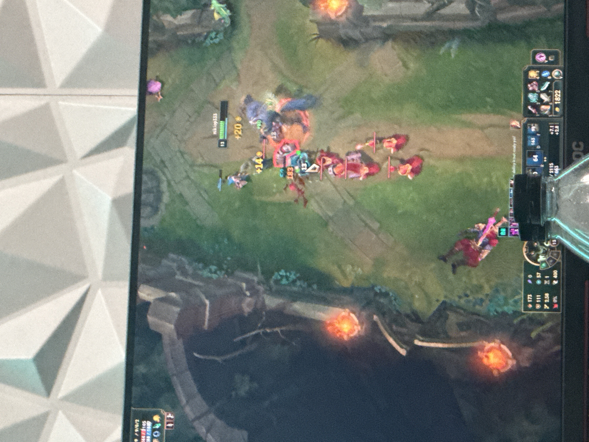 Here is a description of the image:
The image shows a screen capture of a League of Legends match, likely taken with a mobile device. The game is in progress, showing champion characters engaged in a battle. The screen displays game elements such as player names (e.g., "nikiop333"), character levels, health bars, items, and ability cooldown timers. The game interface occupies a significant portion of the image, and parts of the user's environment are visible at the edges of the frame, like a patterned wall and a transparent container with liquid.