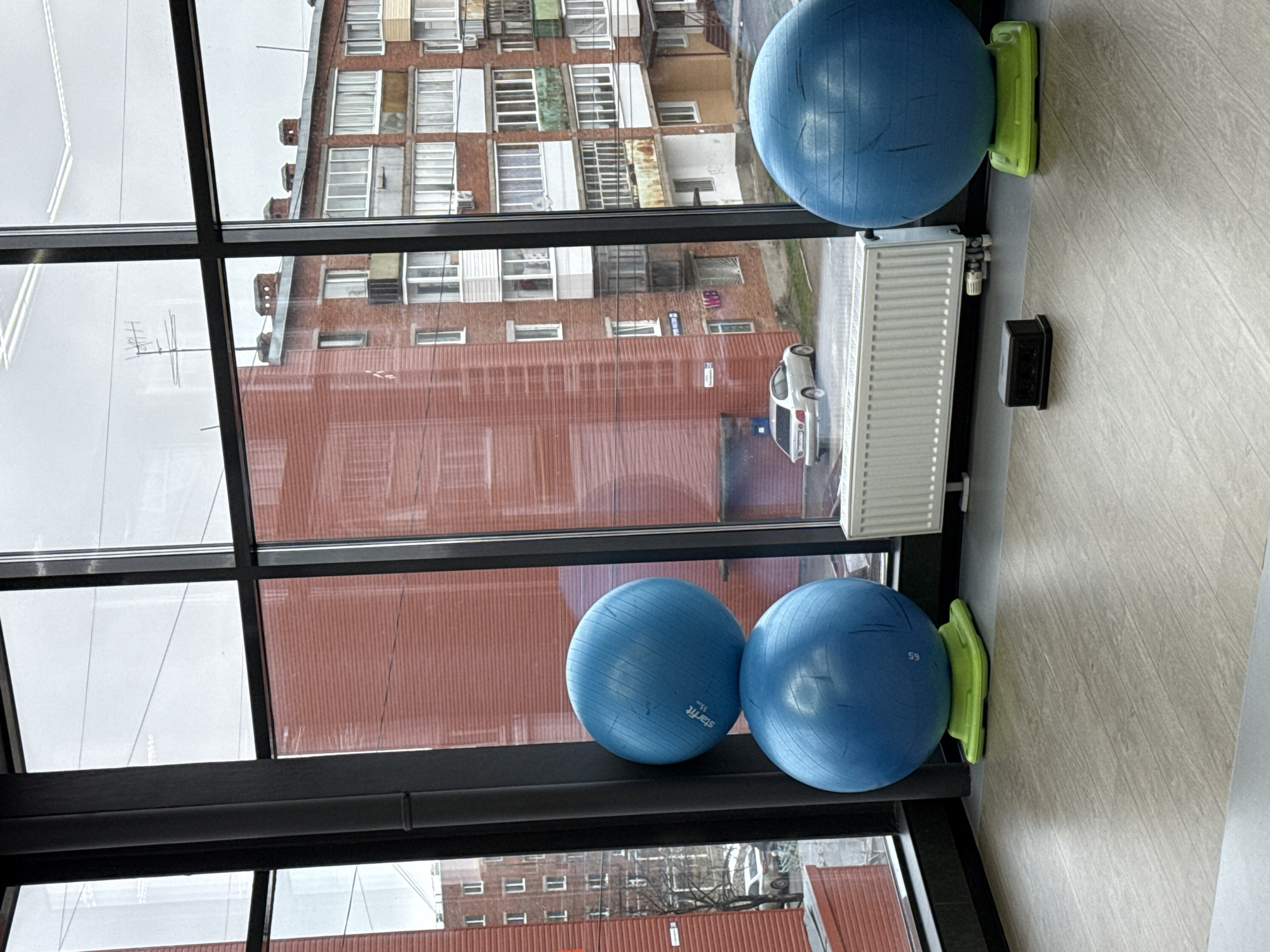 The image shows the interior of a room with a large window providing a view of an urban landscape. Two blue exercise balls are visible inside the room, one at the top of the window and two near the bottom. These balls are placed on what appears to be a window sill. A radiator and a small black rectangular object are positioned near the top exercise ball. The window's frame is dark, contrasting with the outdoor view.
The view through the window reveals buildings of a residential complex. A white car is parked outside in front of the buidlings. The buildings appear to be a multi-story residential building, possibly in Votkinsk, Russia.
