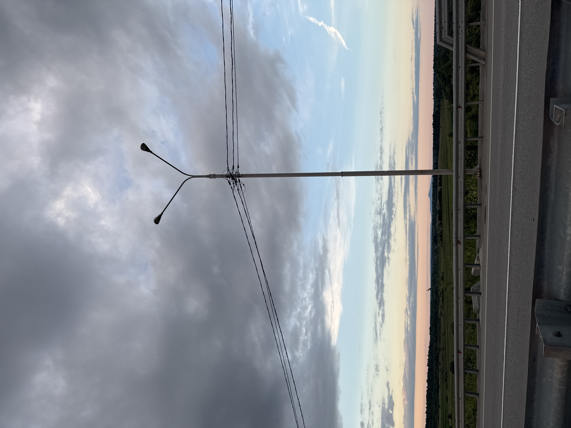 The image depicts a roadside scene at dusk, likely in the vicinity of Ryabinino, Russia. A tall, light pole with two lamps stands prominently in the center, with power lines stretching horizontally across the frame. Below the pole, a highway barrier runs along the edge of the road, which is paved and has a white lane marking. The background features a rural landscape with fields, trees, and a distant hill or mountain range under a cloudy, pastel-colored sky. The overall impression is one of a tranquil, late-day setting in a rural or suburban area.