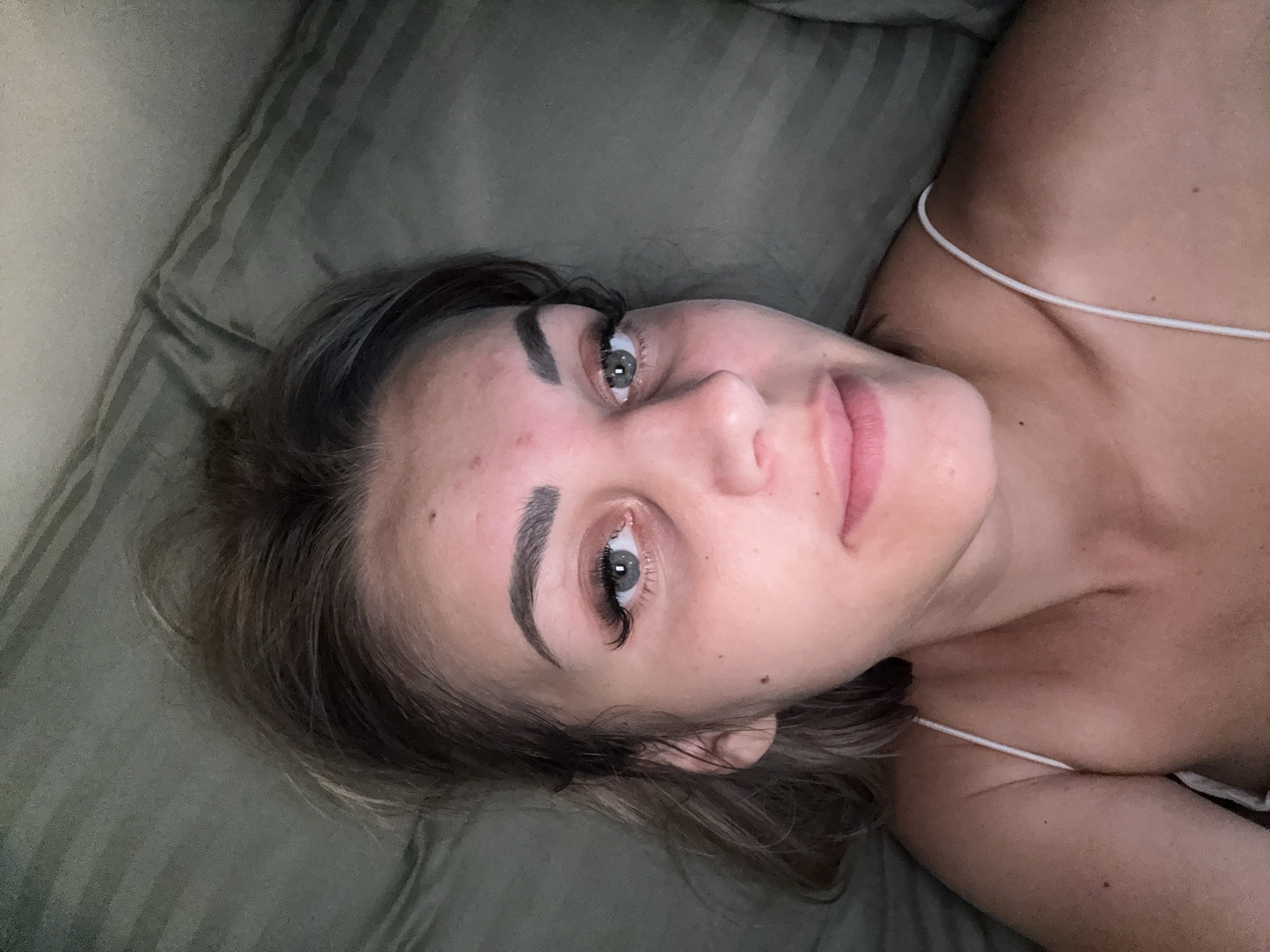The image is an unrotated, eye-level, close-up shot of a young Caucasian woman lying on her side on a textured green surface. She has fair skin, dark blonde hair, and grey eyes. Her head is tilted towards the camera, and her gaze is directed slightly to the right. She is wearing a white spaghetti strap top. The background is a blurry, textured green surface that appears to be a couch or bed. There are no discernible landmarks or environmental details that would place this image specifically in Yuzhno-Sakhalinsk, Russia.