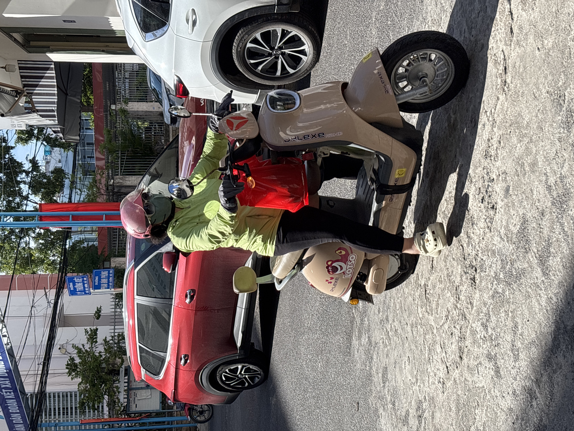 In Nha Trang, Vietnam, a person wearing a green jacket, black pants, a pink helmet, and a green face mask rides a beige electric scooter. They are wearing light-colored Crocs with charms. The scooter has a red bag hanging from the handlebars and a graphic of the character Lotso from Toy Story on the rear panel. The scooter is on a paved road with visible cracks and a shadow cast from the scooter. In the background, a red SUV is parked, and a white SUV is to the right of the scooter. Overhead, power lines crisscross the sky. Signs are visible on a building in the background, including one with Vietnamese text that translates to "Đường Lý Thường Kiệt" which means Ly Thuong Kiet Street, and another that reads "Đường Phan Đình Phùng" meaning Phan Dinh Phung Street.