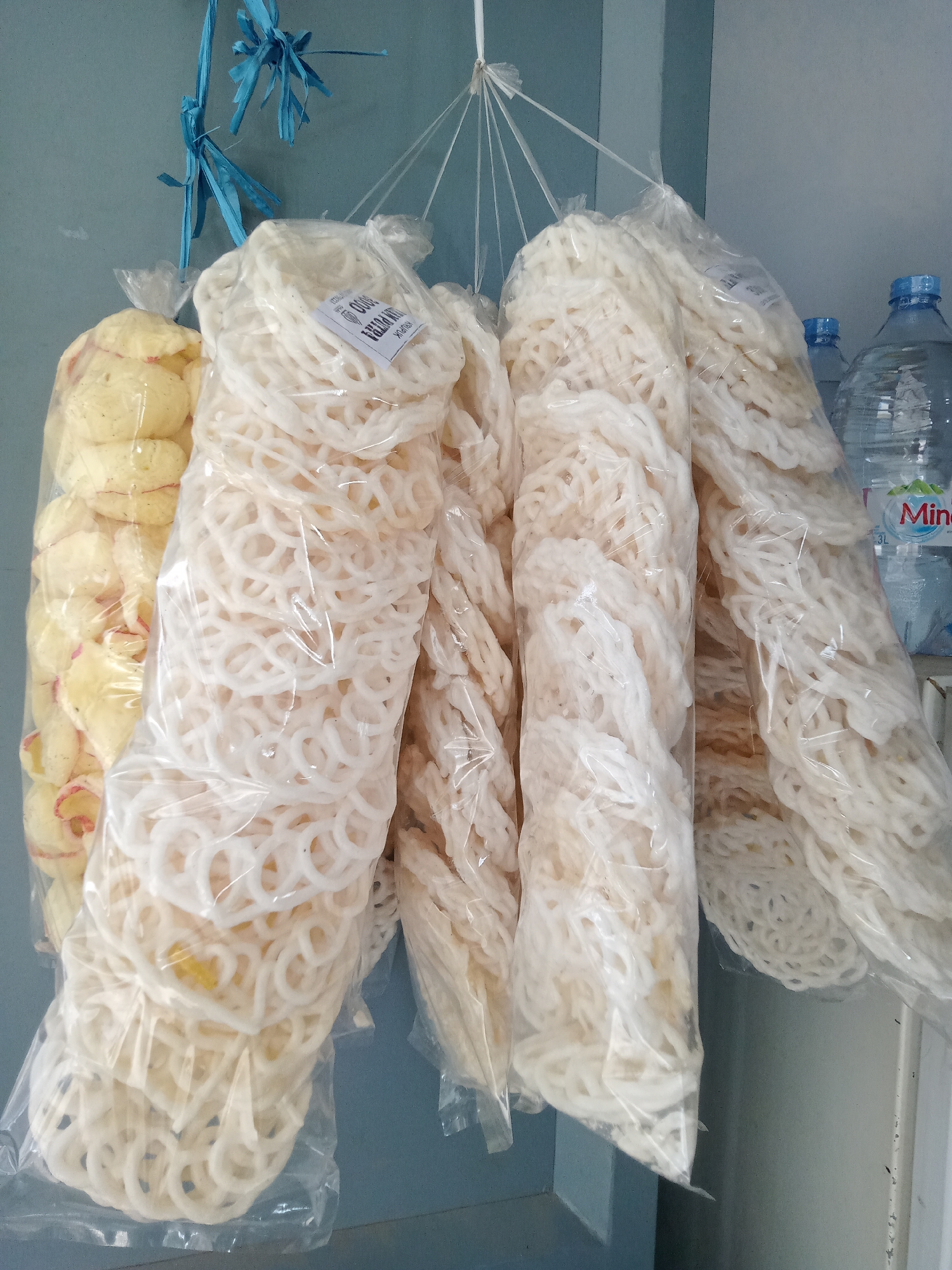 A medium shot captures a display of packaged snacks hanging from a blue plastic string against a muted blue wall. The snacks appear to be a type of white cracker, possibly kerupuk, which are common in Indonesia. There are at least five such packages visible, some filled with circular, ring-shaped crackers and others with what seem to be larger, flattened pieces. To the left of the crackers, a bag filled with yellowish, slightly reddish-pink snack pieces is visible. In the background, partially obscured by the hanging snacks, are two large plastic water bottles, one labeled "Minda". The scene is well-lit, suggesting it is daytime. The overall impression is of a small market stall or a shop display in Simo, Indonesia.