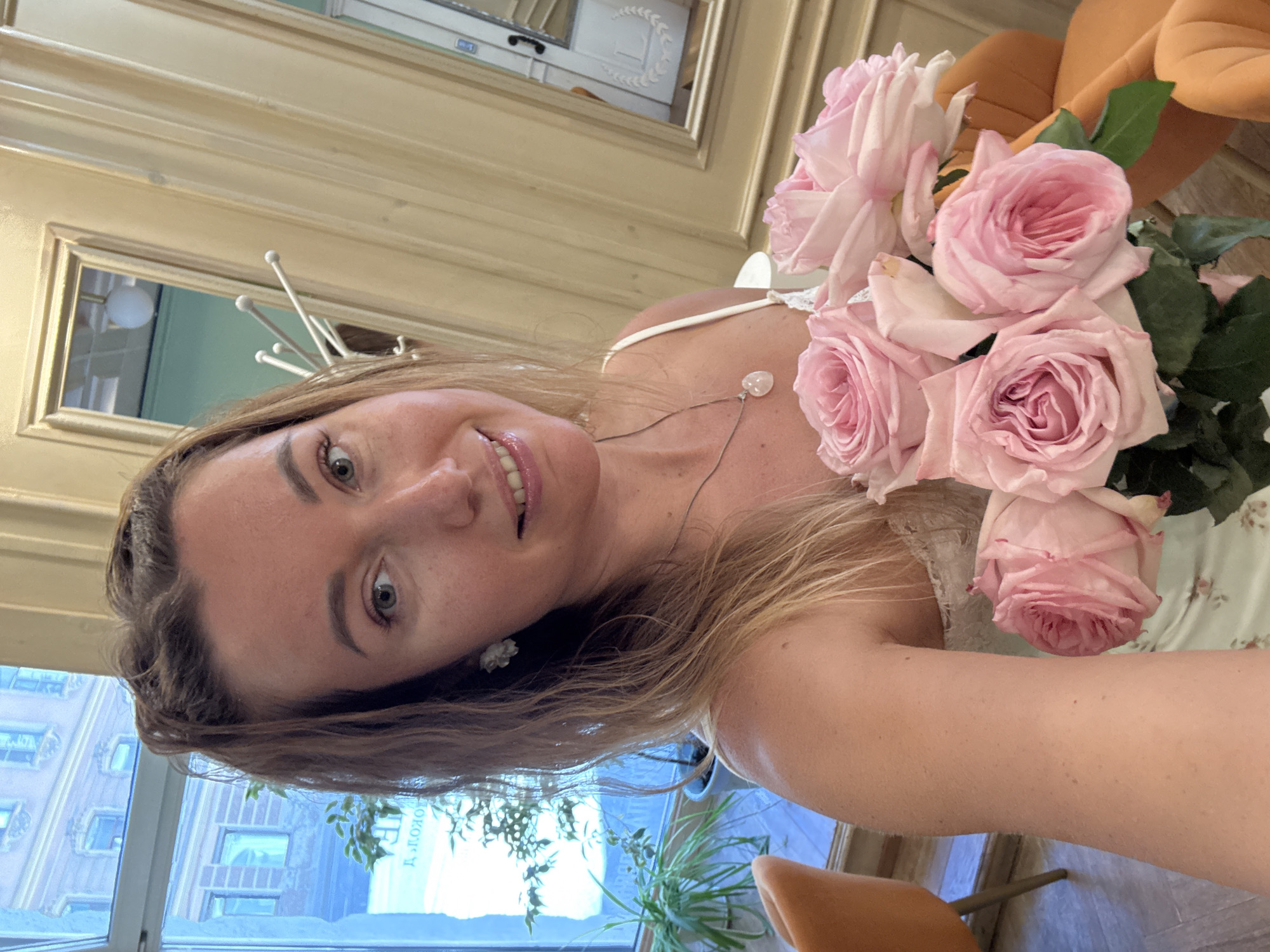A female subject, approximately 20-30 years old, with light brown hair and a fair complexion, is depicted from the chest up, taking a selfie. She has light makeup, including eyeliner and lip gloss, and is smiling slightly. Her left ear (viewer's right) features a white, flower-shaped earring. A silver necklace with a clear or milky heart-shaped pendant rests on her décolletage. She wears a light-colored, possibly white, strappy top with lace trim. In her left arm (viewer's right), she holds a bouquet of five to six light pink roses with green foliage.
The background indicates an indoor setting, likely a cafe or restaurant, in an urban environment in an unknown city. A large window on the left reveals an exterior view of a multi-story building with multiple windows across the street, featuring classic architectural details. Partially visible text, appearing as "Д.ПОКОЛО" in reversed Cyrillic script, is reflected on the window pane. Potted plants are positioned near the window. The interior features light-colored paneled walls with framed mirrors or decorative panels on the right side. One mirror reflects a white, multi-hook coat rack and a spherical light fixture. Another reflection shows a white door with a dark handle and a subtle logo or emblem. Two orange upholstered chairs are visible in the background, one in the lower left corner and another on the right side. The floor appears light-colored, possibly wood or tile.
