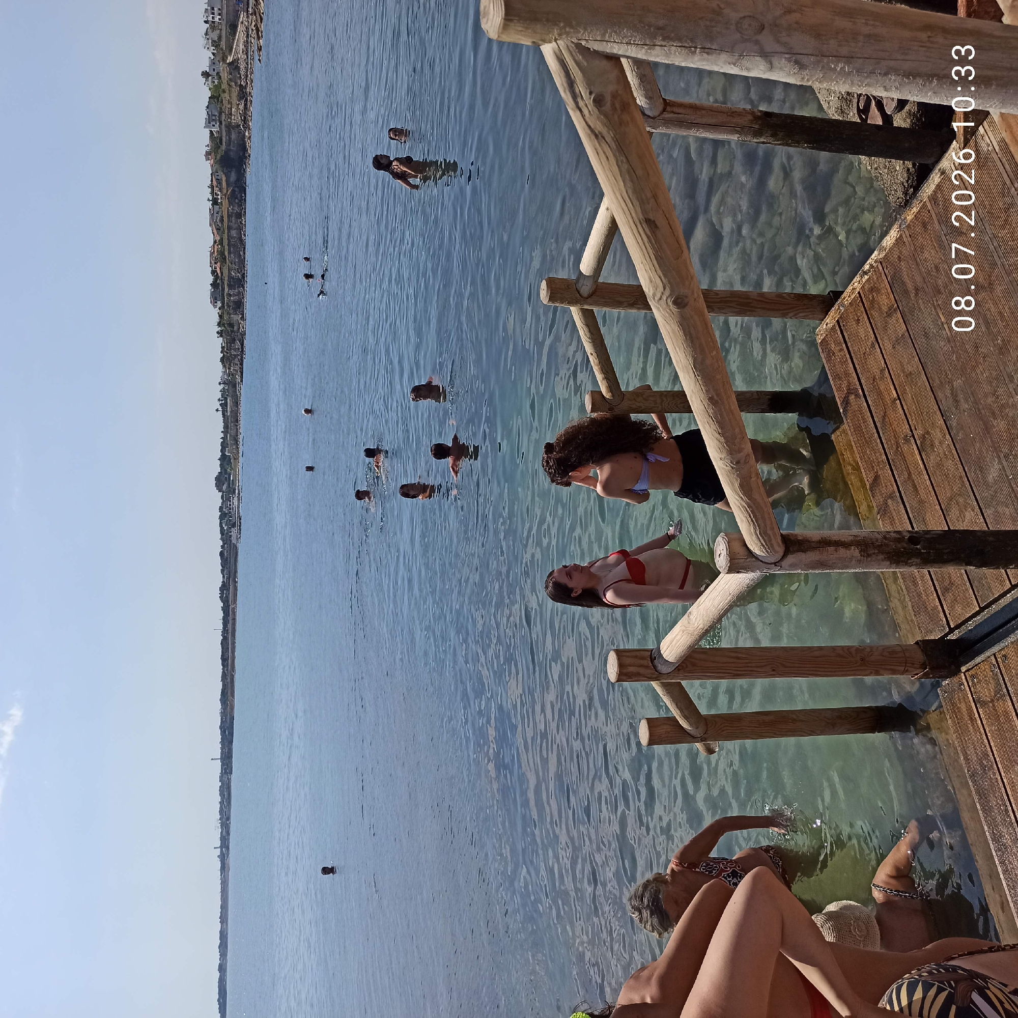 In Trani, Italy, on July 8, 2026, at 10:33 AM, a group of people are in the clear, shallow water of the sea. A wooden walkway with railings leads down to the water. In the foreground, two young women are standing in the water, facing each other. One is wearing a red bikini, and the other has a light blue bikini top and dark shorts. Behind them, several other individuals are swimming or wading further out in the water. In the distance, a coastline with buildings and vegetation is visible under a clear blue sky. The seabed is visible through the transparent water, with rocks and sand.