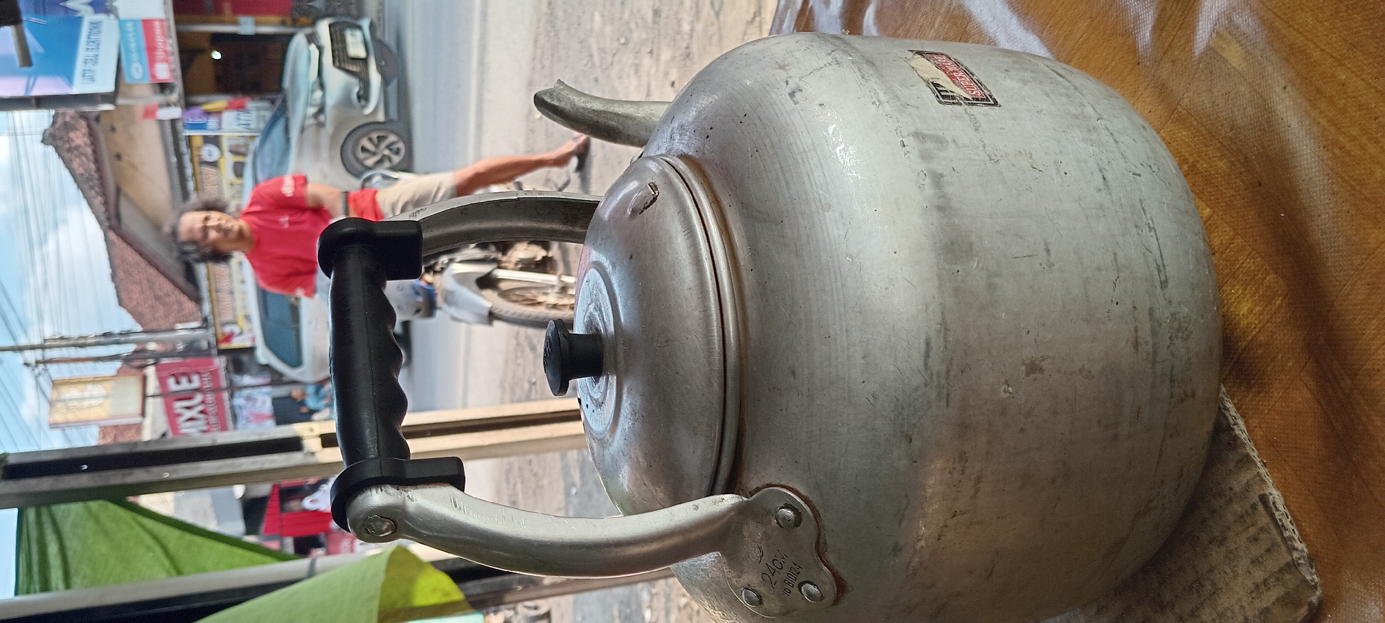 In Karangsari, Indonesia, a close-up shot focuses on a dented, aluminum kettle with a black handle and a black lid knob. The kettle rests on a textured surface, possibly a woven mat or tablecloth, with a piece of corrugated cardboard underneath. In the blurred background, a man in a red t-shirt and light-colored shorts is visible, standing near a silver car and a motorcycle. Behind him, buildings with tiled roofs and street signs, including one that reads "MIXLE," indicate an urban or semi-urban setting. The overall impression is of a scene from an outdoor roadside stall or eatery.