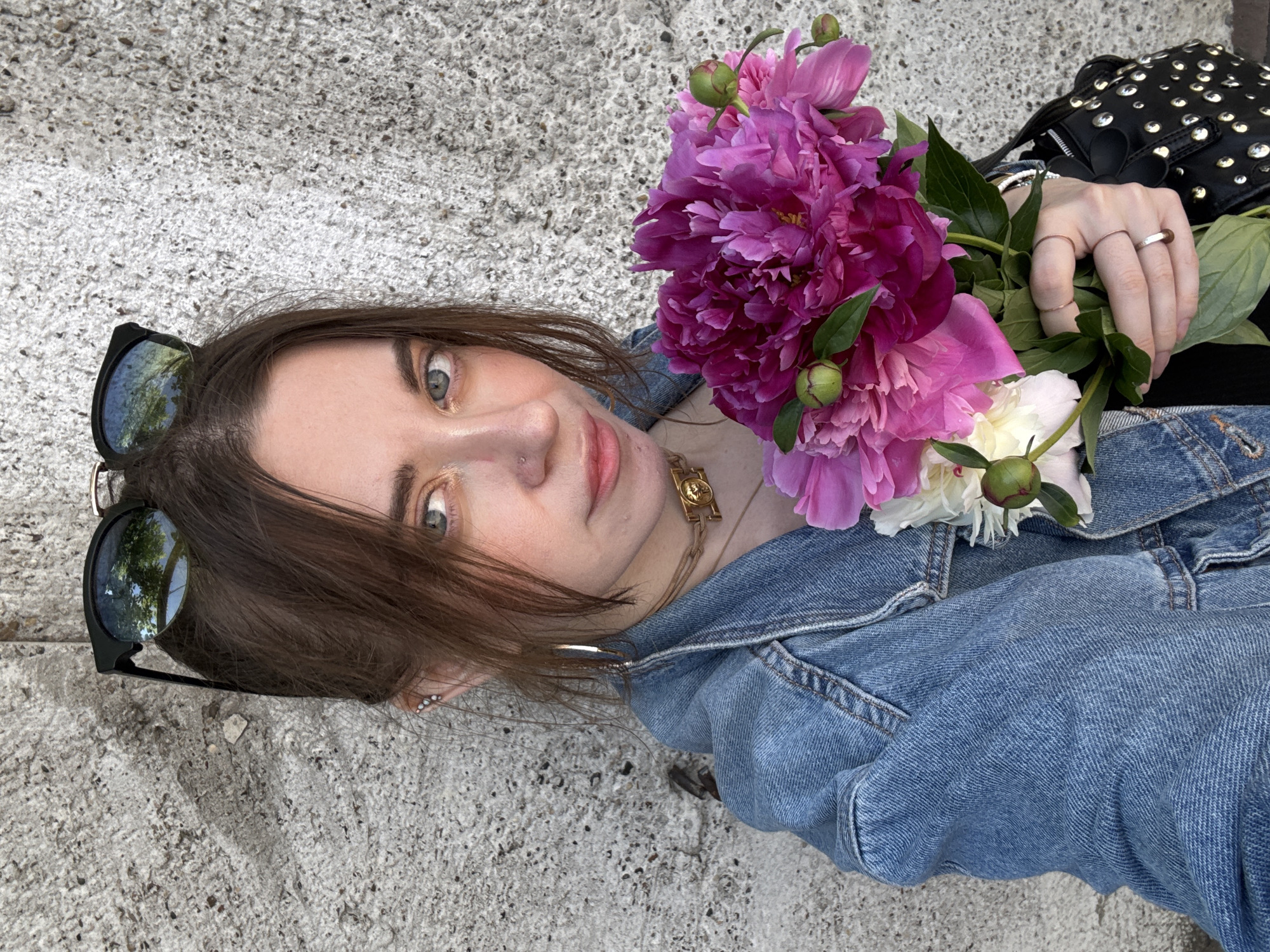 A young Caucasian woman is pictured outdoors, holding a bouquet of pink and white peonies. She is wearing a denim jacket, a gold necklace, and several rings on her fingers. Sunglasses are perched on top of her head. The background is a textured, gray concrete wall. The woman's left eye is visible, and she is looking towards the camera. Her hair is dark brown and pulled back. The lighting suggests it is daytime.