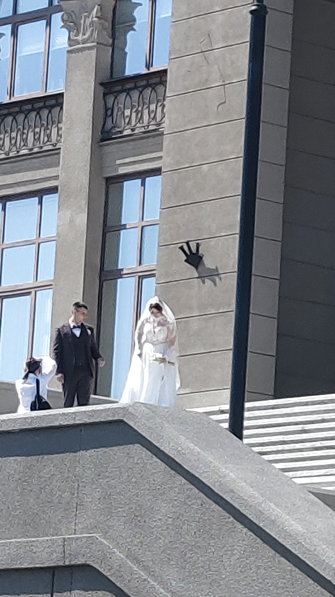 This outdoor photograph captures a wedding photoshoot featuring a bride and groom, along with a partially visible third individual, possibly a photographer, assistant, or guest. The scene is set on a grand, multi-tiered grey stone staircase or platform, leading up to a large, imposing building.
The groom, positioned slightly to the left, wears a dark suit, a white shirt, and a black bow tie. He is looking towards the bride. The bride stands to the right of the groom, dressed in a traditional white wedding gown with a flowing veil. She holds a small, white floral bouquet in her hands. The third person is only partially visible on the far left, appearing to wear a white top and dark lower garment.
The backdrop is a multi-story building constructed from grey stone or concrete, featuring large, rectangular windows with light blue reflections, indicating a clear sky. Several windows on an upper floor are adorned with ornate, dark iron balconies. A striking black, crown-like sculpture is mounted on the building wall to the right of the couple, casting a prominent shadow below it. A dark lamppost or utility pole runs vertically on the far right of the frame.
The image appears to be taken on a bright, sunny day, evidenced by the strong shadows cast by the sculpture and the overall clear lighting. No visible text can be discerned in the image. The architectural style and the presence of the crown sculpture, given the location context of Krasnoyarsk, Russia, suggest the couple is posing at a significant landmark or public building in the city.