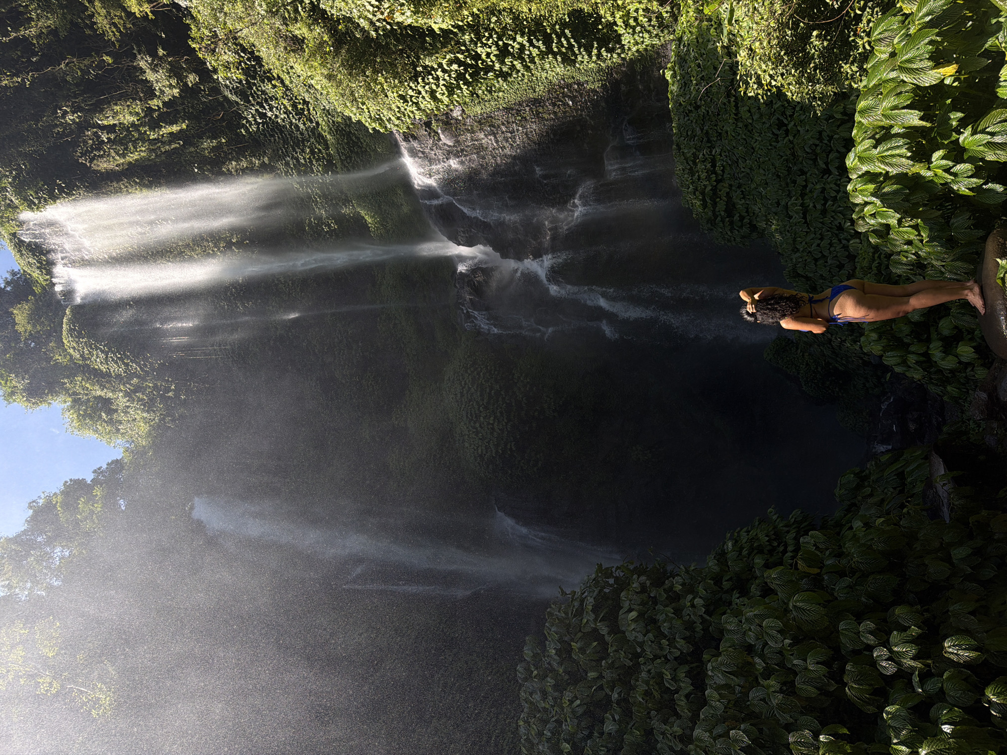 In the city of Sawan, Indonesia, a woman stands on a rock at the base of a cascading waterfall. She is wearing a blue bikini, and her back is to the camera as she looks up at the waterfall. Lush green foliage surrounds the waterfall and the woman, with sunlight filtering through the trees and mist. The waterfall itself is tall and multi-tiered, with water flowing down a rocky cliff face. The overall scene depicts a serene natural landscape with a solitary human presence.