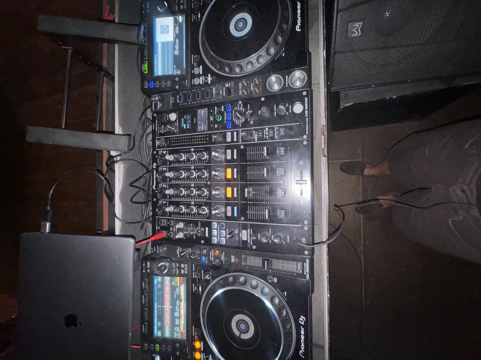 The image shows a DJ setup with two Pioneer CDJ-2000NXS2 media players, a Pioneer DJM-900NXS2 mixer, and a MacBook laptop connected to the equipment with cables. A speaker marked with a "3" is visible in the upper right corner. The setting appears to be indoors with dark lighting. There are feet that suggest an adult standing next to the sound board. There is no evidence to support that this image was taken in Moscow, Russia.