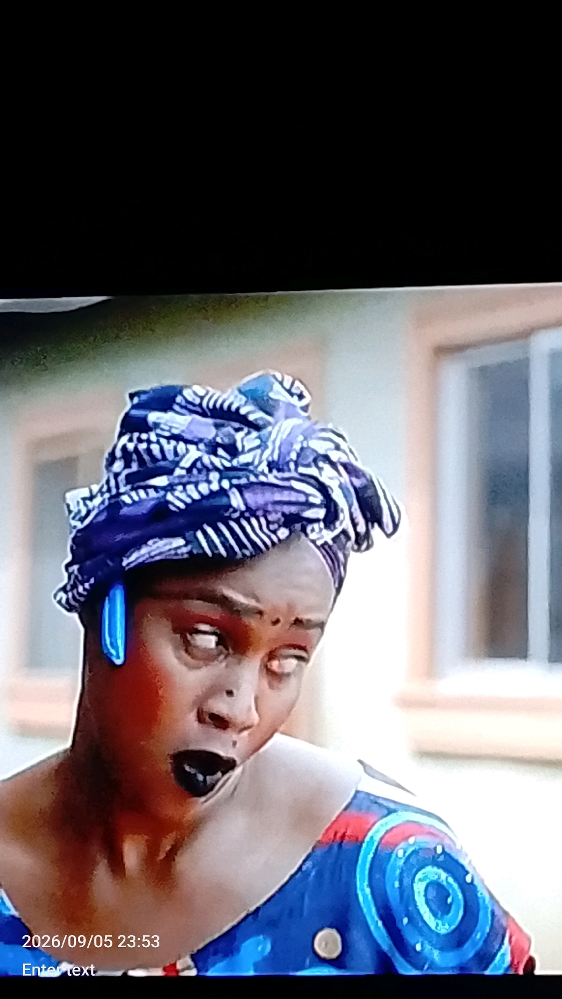A woman with dark skin is shown from the chest up, wearing a vibrant blue and red patterned dress and a blue and white patterned headwrap. She has bright blue earrings and dark lipstick. Her eyes are rolled back, and her mouth is open. A timestamp in the lower left corner reads "2026/09/05 23:53" and an "Enter text" prompt is below it. The background appears to be a building with windows, suggesting an outdoor setting, possibly in Lokoja, Nigeria, given the provided context.