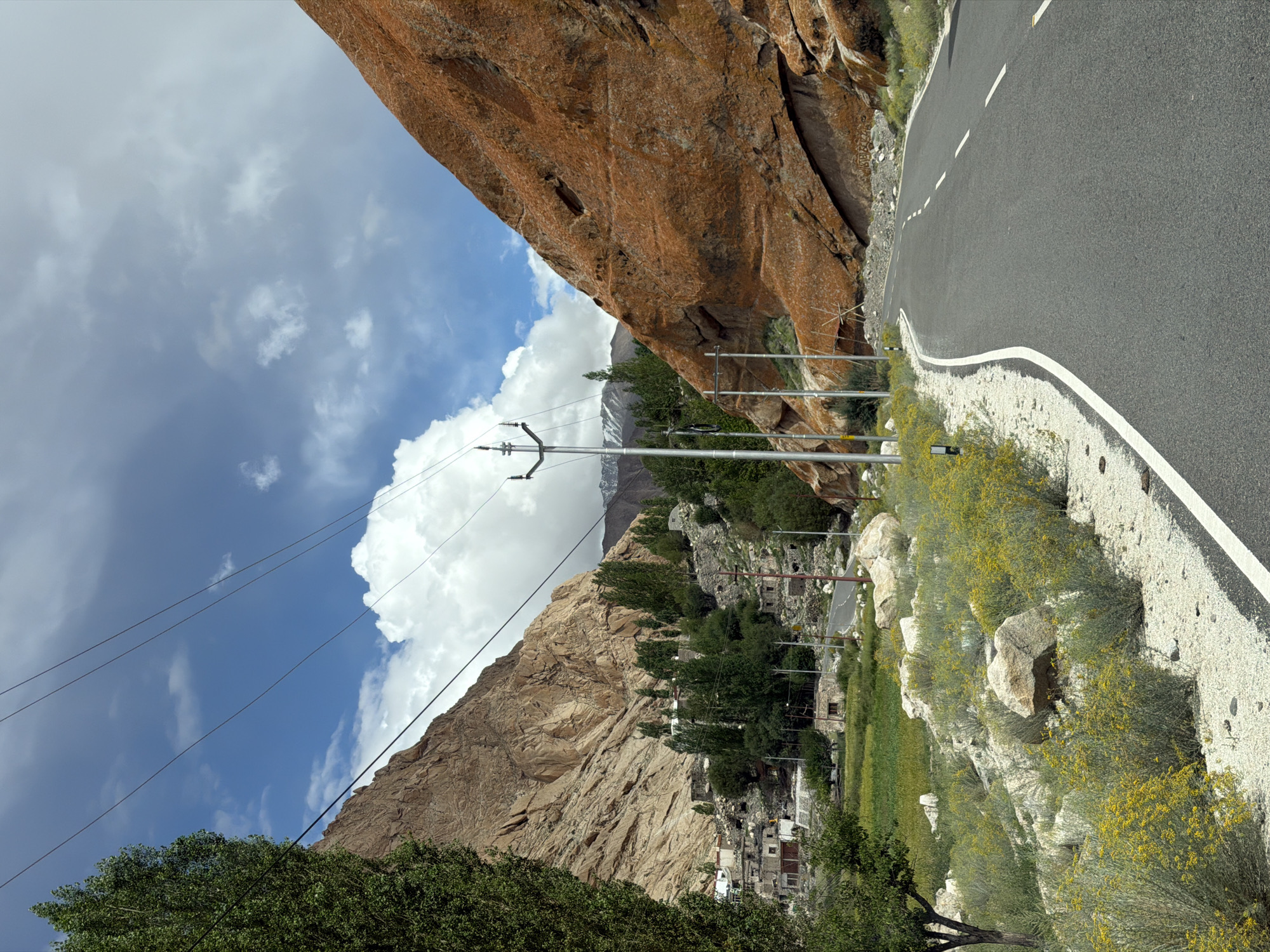 A winding asphalt road cuts through a mountainous landscape under a partly cloudy sky. The road, bordered by white lines, curves to the right. On the left, a steep, rocky mountain face rises, covered in sparse green vegetation at its base. A tall, leafy green tree stands in the foreground on the left. Further back, nestled in the valley, a small village with stone houses and scattered trees is visible. The right side of the road is dominated by a massive, orange-colored rock formation, textured and uneven. Power lines stretch across the frame, supported by utility poles. The ground along the roadside is covered in gravel and dotted with small yellow wildflowers. In the distance, snow-capped mountains are faintly visible. The location is identified as Hymia, India.