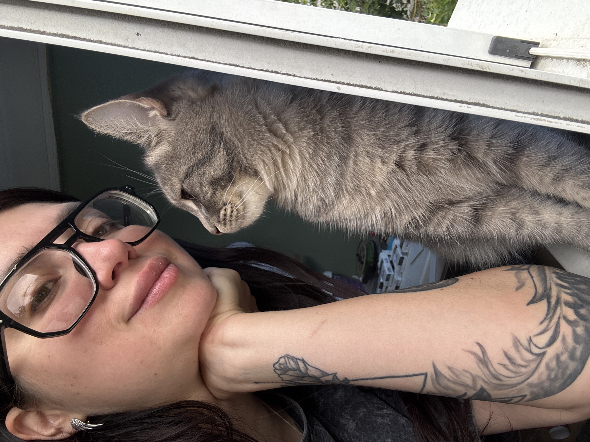 A woman is in the foreground, looking to her left, with her chin resting on her hand. She is wearing large, black-rimmed glasses and a gray t-shirt. Her left arm is visible, featuring several tattoos. Next to her, also looking to her left, is a gray tabby cat. Both subjects are positioned next to a window frame, with the background showing a green wall and some out-of-focus objects on a surface. The scene appears to be indoors, near a window.