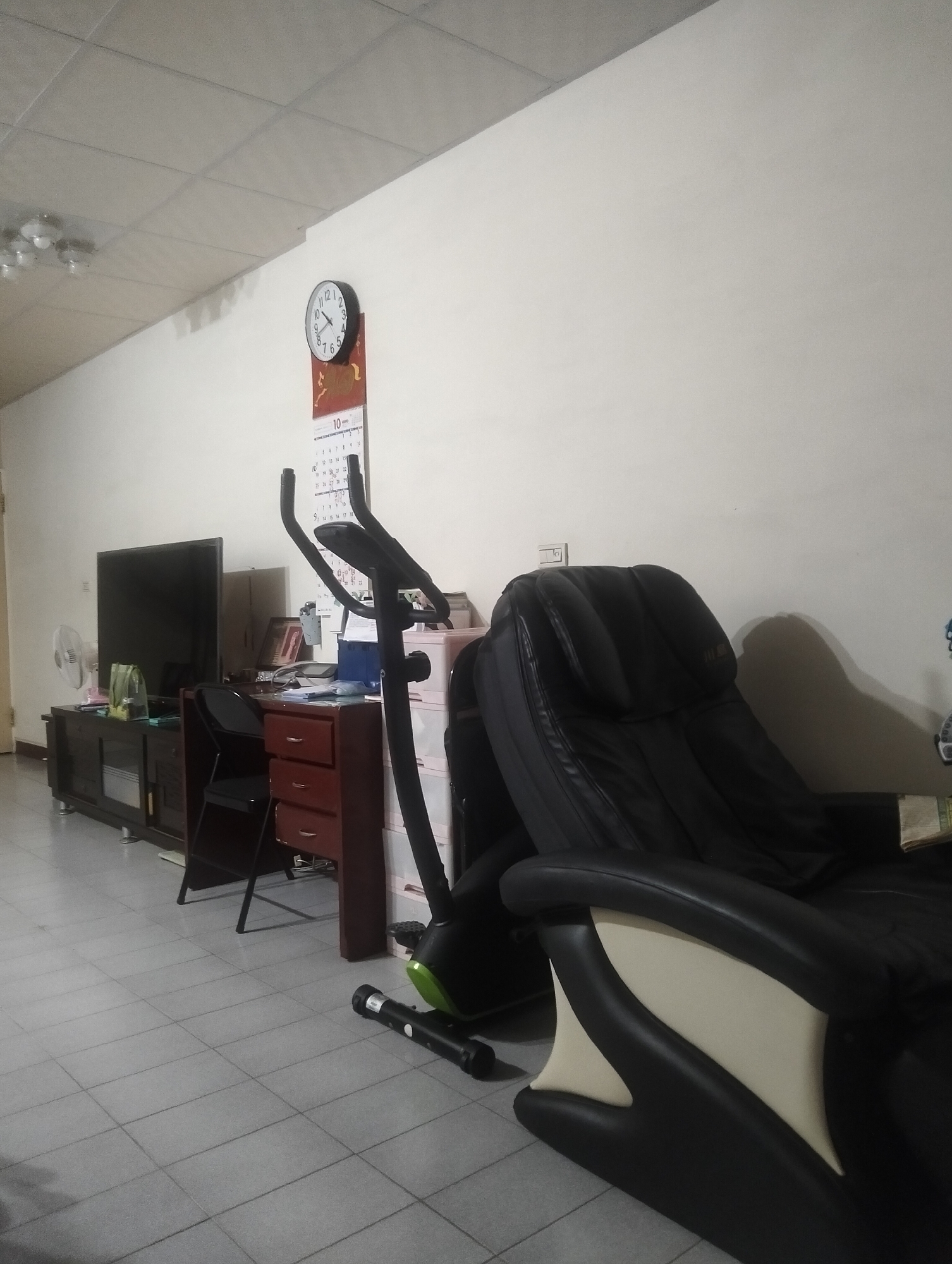 The image depicts an indoor scene within a room, likely a living area or study, in Kaohsiung, Taiwan. The room is furnished with various items. Dominating the right side of the frame is a large, black leather massage chair with cream-colored accents. To the left, a dark wooden entertainment console supports a large, black television. Beside the console, a smaller, dark wooden desk with red drawers is visible, with a black folding chair placed in front of it. An exercise bike, mostly black with some green accents, stands behind the desk and to the right of the television. A white oscillating fan is positioned on the floor to the left of the entertainment console. A round wall clock with a white face and black numerals is mounted on the wall above a calendar. The walls are painted white, and the floor is tiled with large, light gray square tiles. The ceiling appears to be a dropped ceiling with fluorescent lighting fixtures, some of which are partially visible. The overall impression is of a functional, lived-in domestic space.
