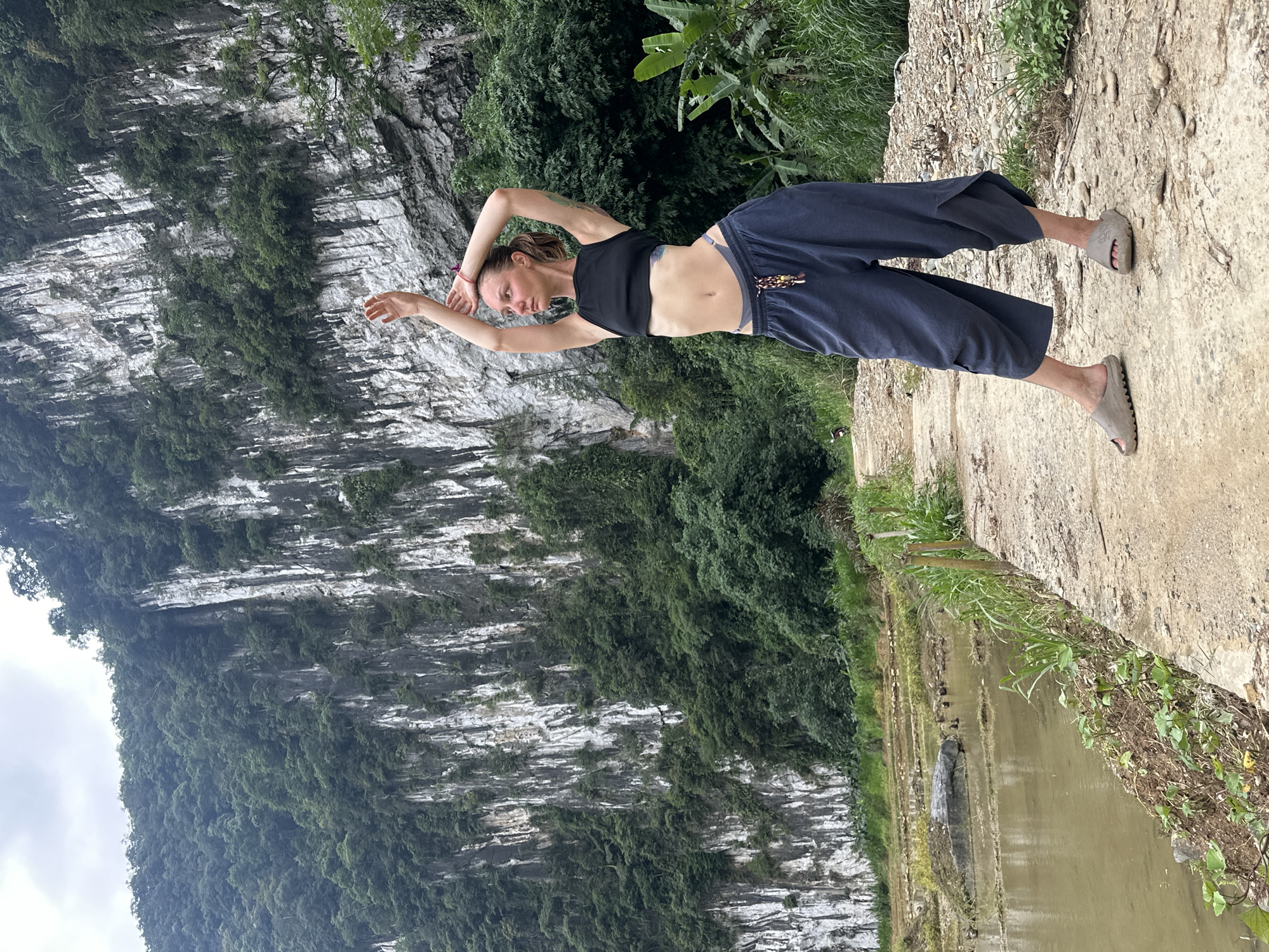 A young woman with blonde hair pulled into a ponytail, wearing a black crop top, grey capri pants, and grey sandals, poses with her arms raised above her head on a dirt path. She is standing in front of a steep, rocky cliff face covered in green foliage. To her left is a flooded rice paddy with a few small birds visible. The scene is set in the Pù Luông Commune in Vietnam, characterized by its dramatic karst landscape.