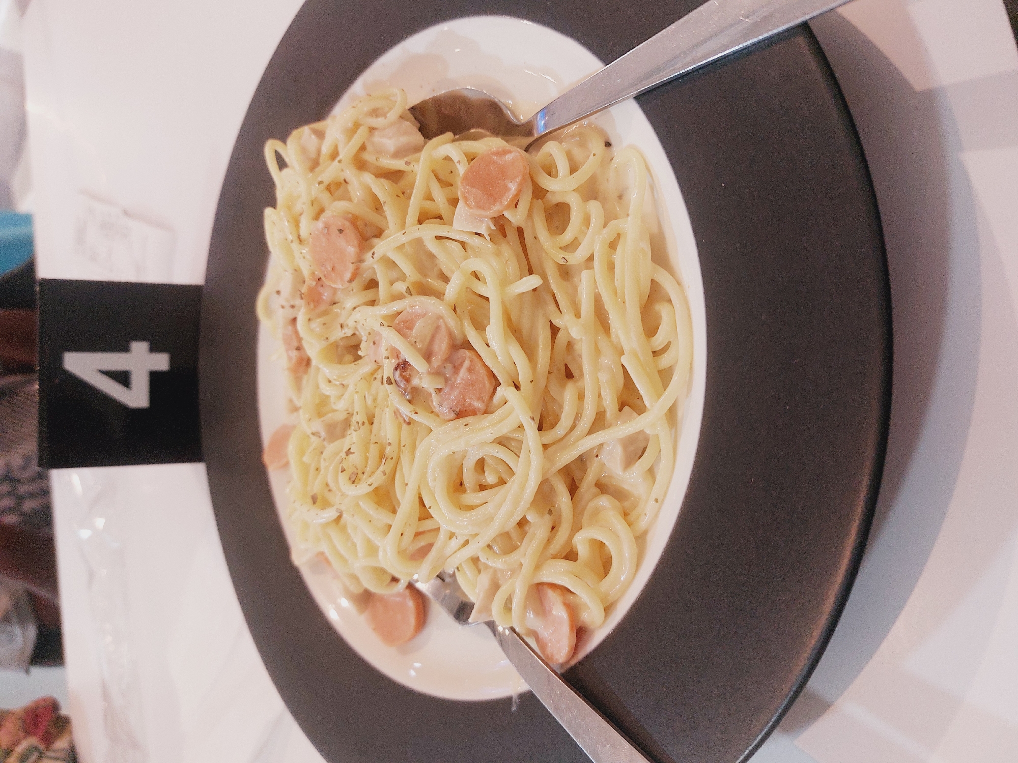 The image shows a close-up of a plate of spaghetti carbonara, garnished with herbs. Two silver utensils, a fork and a spoon, are placed on either side of the plate, suggesting the meal is ready to be eaten. In the background, a black number sign with a white numeral '4' is visible, possibly indicating a table number in a restaurant. The setting appears to be indoors, with a white tablecloth and hints of other patrons blurred in the background. There is no specific indication that this image was taken in Nunukan, Indonesia.