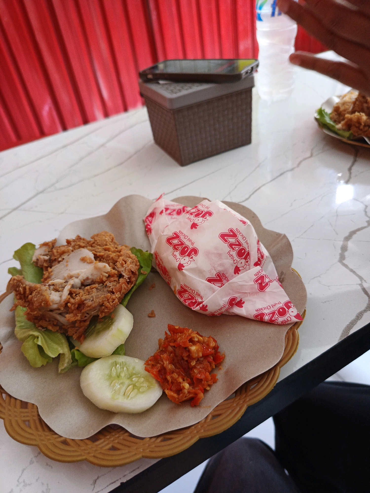 In Tompokersari, Indonesia, a close-up shot captures a table with a meal. The meal consists of fried chicken, sliced cucumbers, and chili paste served on a brown paper wrapper placed in a woven basket. A white plastic bottle of water and a dark brown container with a phone on top are also visible on the white marble table. In the background, red curtains are partially drawn. A blurred human hand is also visible in the upper right corner of the image.