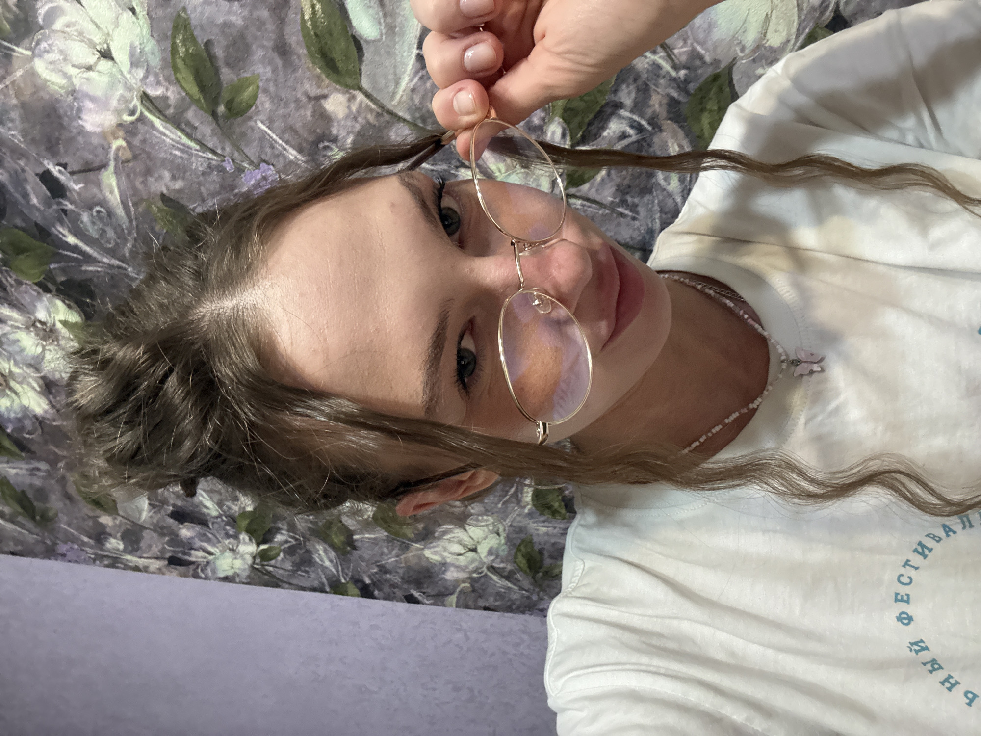 A close-up, front-facing, selfie-style photograph shows a young Caucasian female with light brown hair pulled into a messy bun with strands falling around her face. She is wearing clear, round-framed glasses and a white t-shirt with a light blue, curved inscription on the left chest area. She has a delicate, beaded necklace with a small pink butterfly pendant. Her right hand is lifting the edge of her glasses, and her expression is playful. The background is a floral-patterned wallpaper in muted greens and purples. No specific location in the city of Perm, Russia, can be identified from the image.