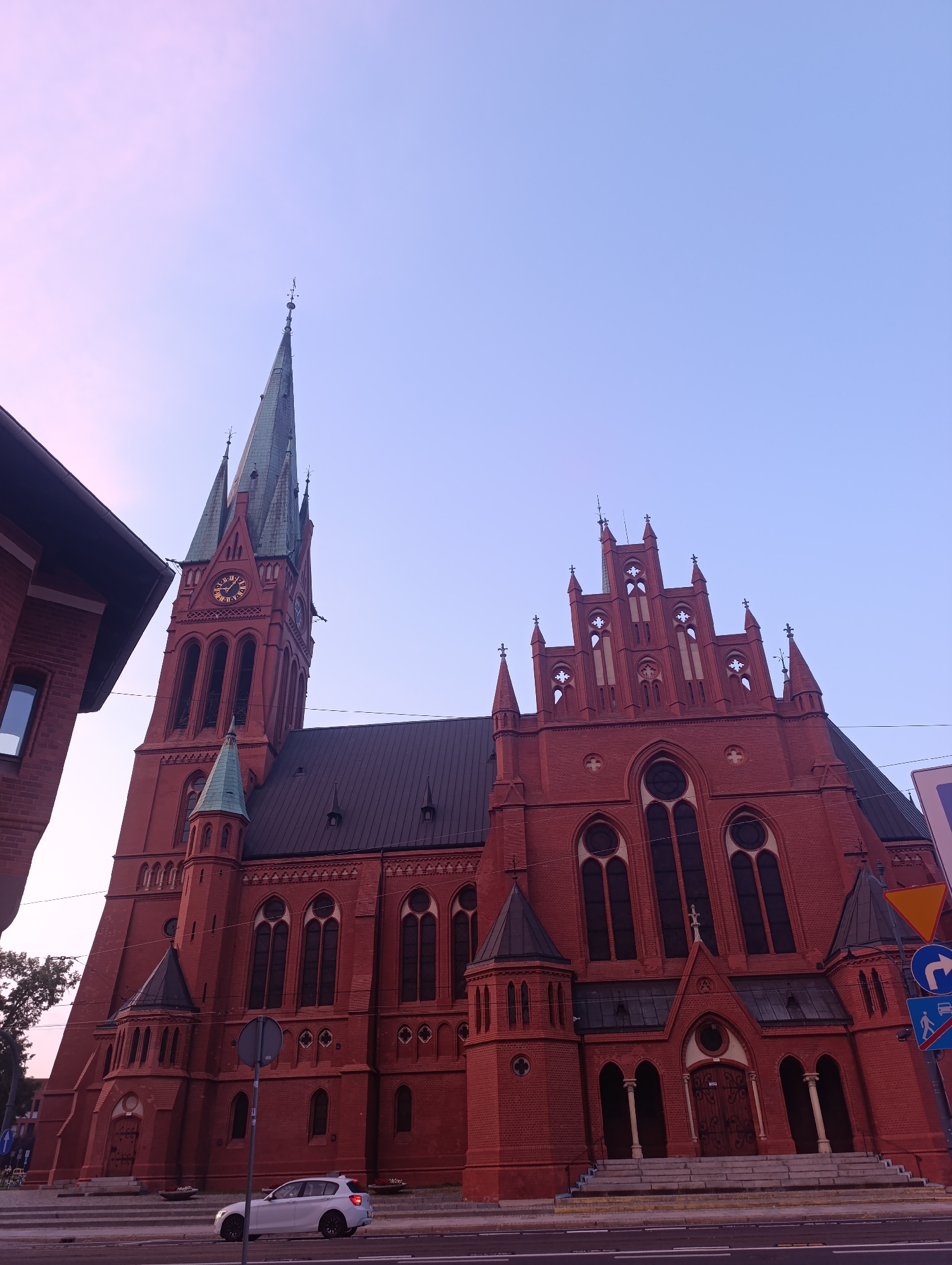 The image depicts the St. James' Church in Toruń, Poland, situated on a street corner. The imposing red brick Gothic Revival church dominates the frame, with its tall spired tower featuring a clock face prominently displayed. The sky above is a gradient of pale pink and light blue, suggesting either dawn or dusk. In the foreground, a white car is parked on the asphalt road, and a pedestrian crossing is visible. A traffic sign indicating a right turn and a pedestrian crossing is also present. The building to the left of the church is a modern brick structure, partially obscuring the view of the church's left side. The overall scene captures the grand architecture of the historic church against a serene sky, with elements of urban life present on the street.