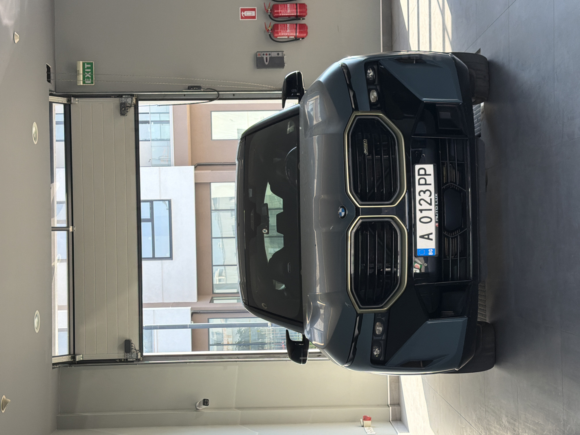 A dark green BMW SUV is parked inside a garage with an open overhead door. The car is facing the camera and its license plate is visible, reading "A 0123 PP BG AUTO LUX". On the wall to the right of the car, there is a green "EXIT" sign, a fire alarm, and two red fire extinguishers. To the left of the car, there is a white security camera mounted on the wall. The garage floor is dark gray tile. In the background, through the open garage door, a modern building with glass windows and balconies can be seen. The lighting in the garage suggests it is daytime. The scene appears to be a car dealership or a similar commercial establishment in Burgas, Bulgaria.